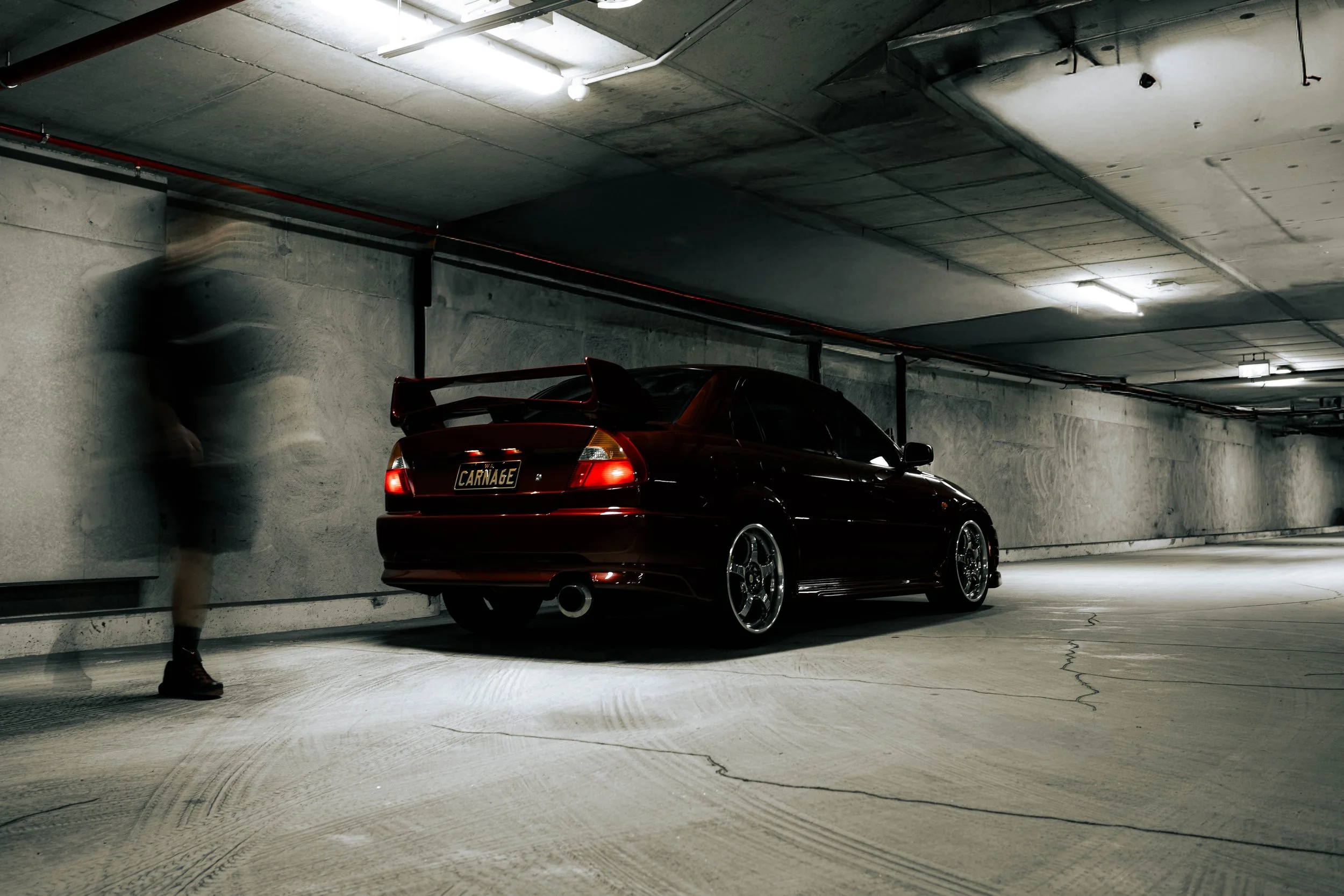 A black sports car with a red spoiler and a custom license plate reading 'CARNAGE' parked in a dimly lit underground parking garage. A blurred person walking past the car.
