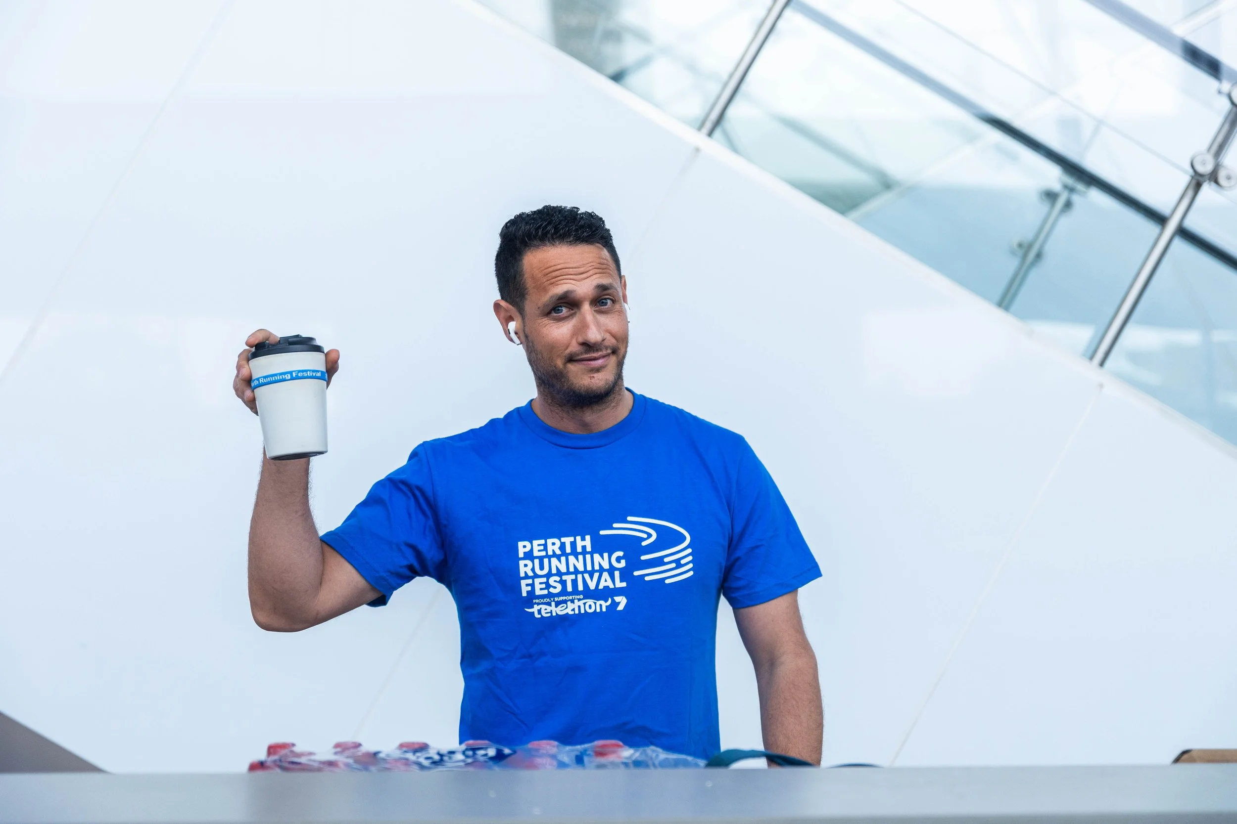 A man in a blue T-shirt holding a coffee tumbler, standing behind a table with promotional items, at the Perth Running Festival.
