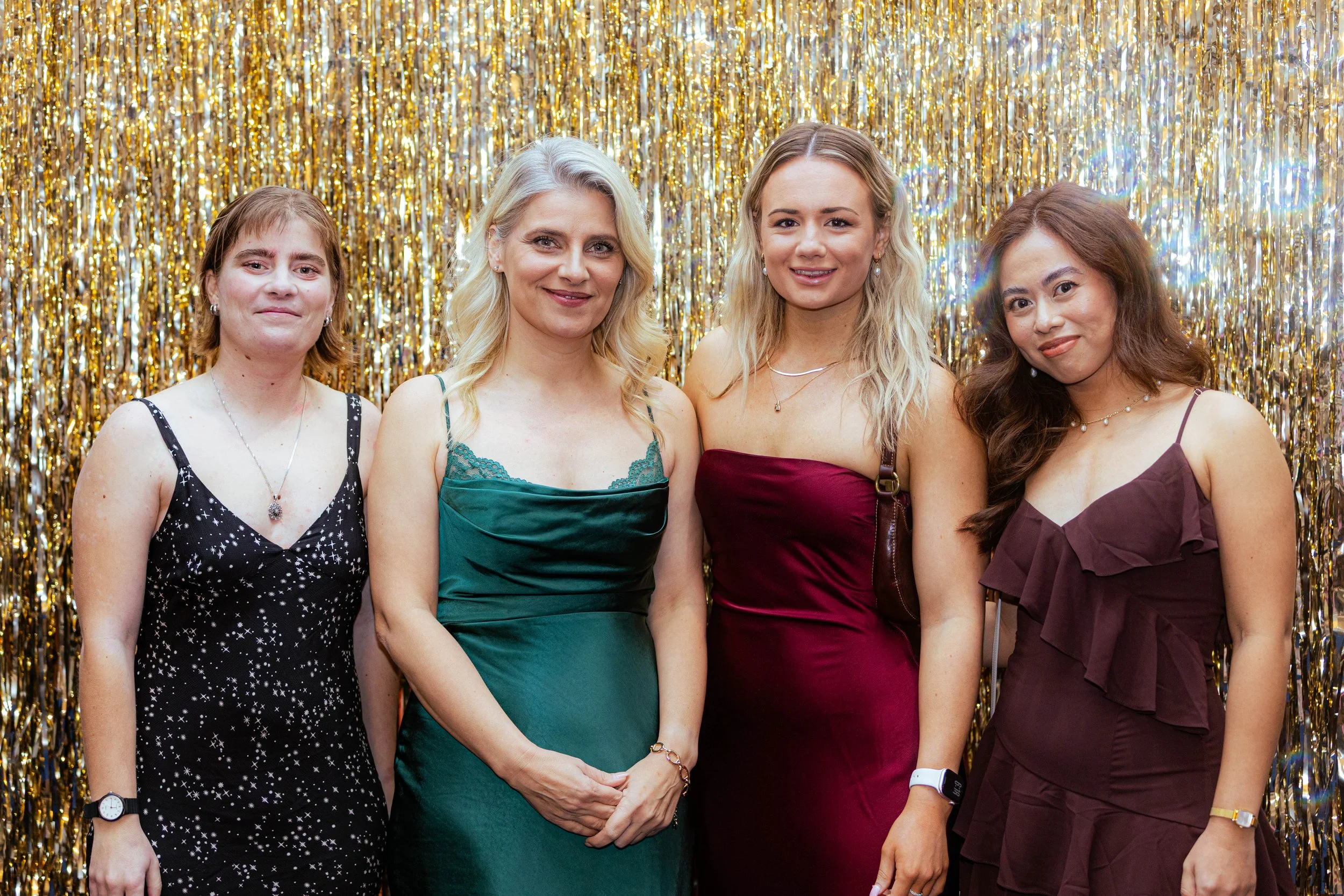 Group of four women standing in front of a gold tinsel backdrop, dressed in semi-formal attire