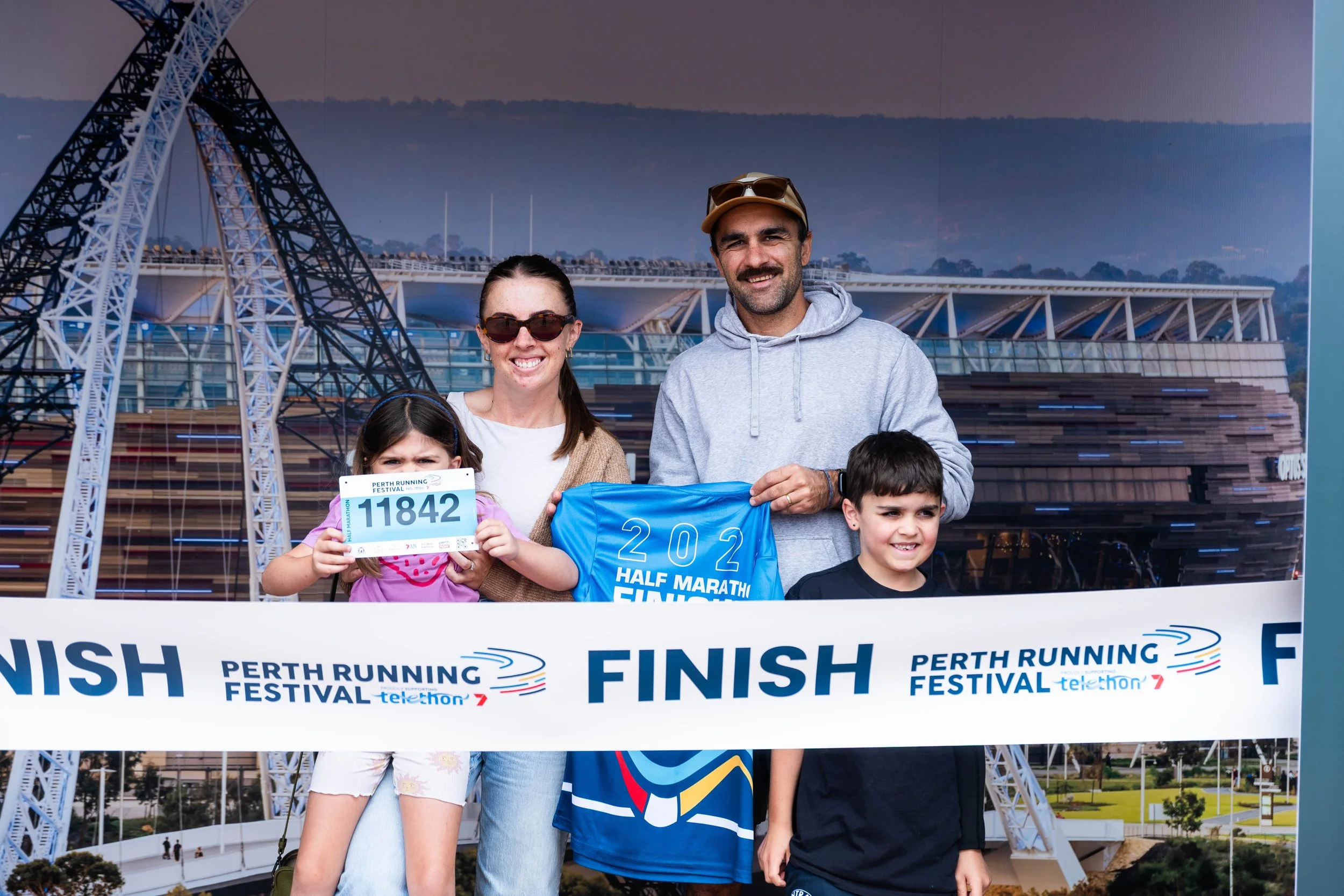 A group of people, including children and adults, standing at the finish line of a marathon event, with a large Ferris wheel and a stadium structure in the background. They are smiling and holding a banner that reads '2022 Half Marathon Finish'.