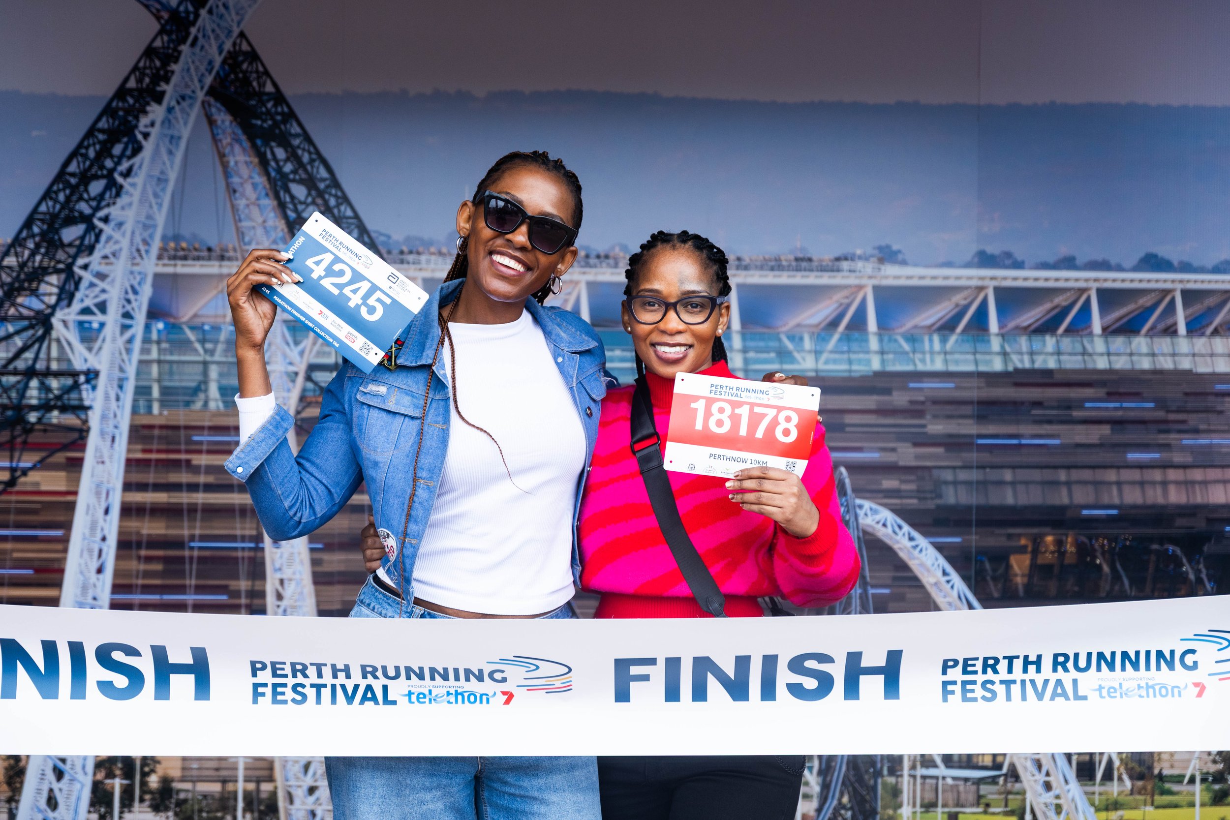 Two women smiling at the finish line of a marathon event, holding race bibs, with the Perth Running Festival banner and a large bridge structure in the background.