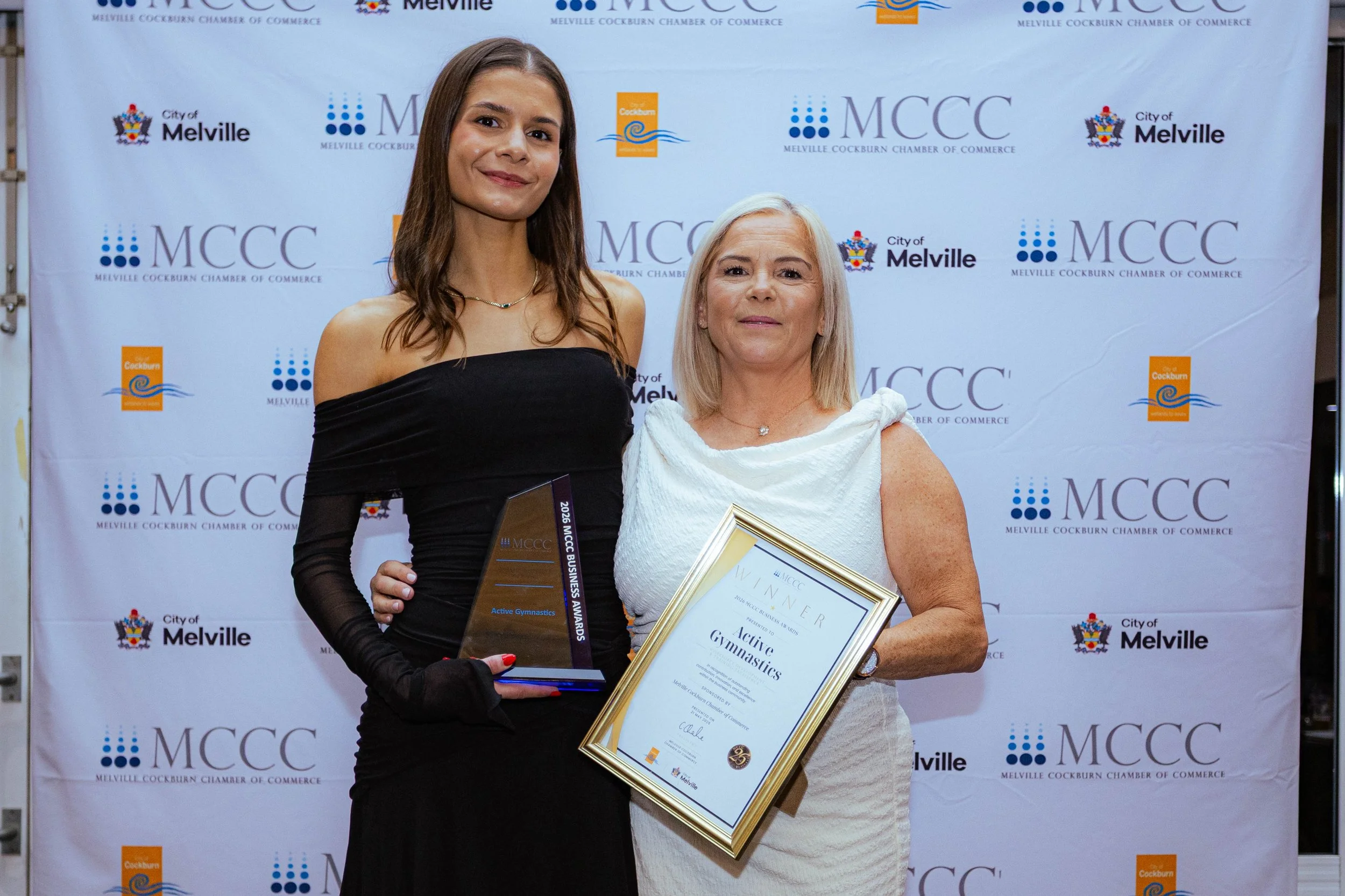 Two women standing in front of a backdrop with logos, one holding an award and the other holding a framed certificate. The woman on the left has long brown hair, is wearing a black off-shoulder dress, and has a necklace. The woman on the right has li