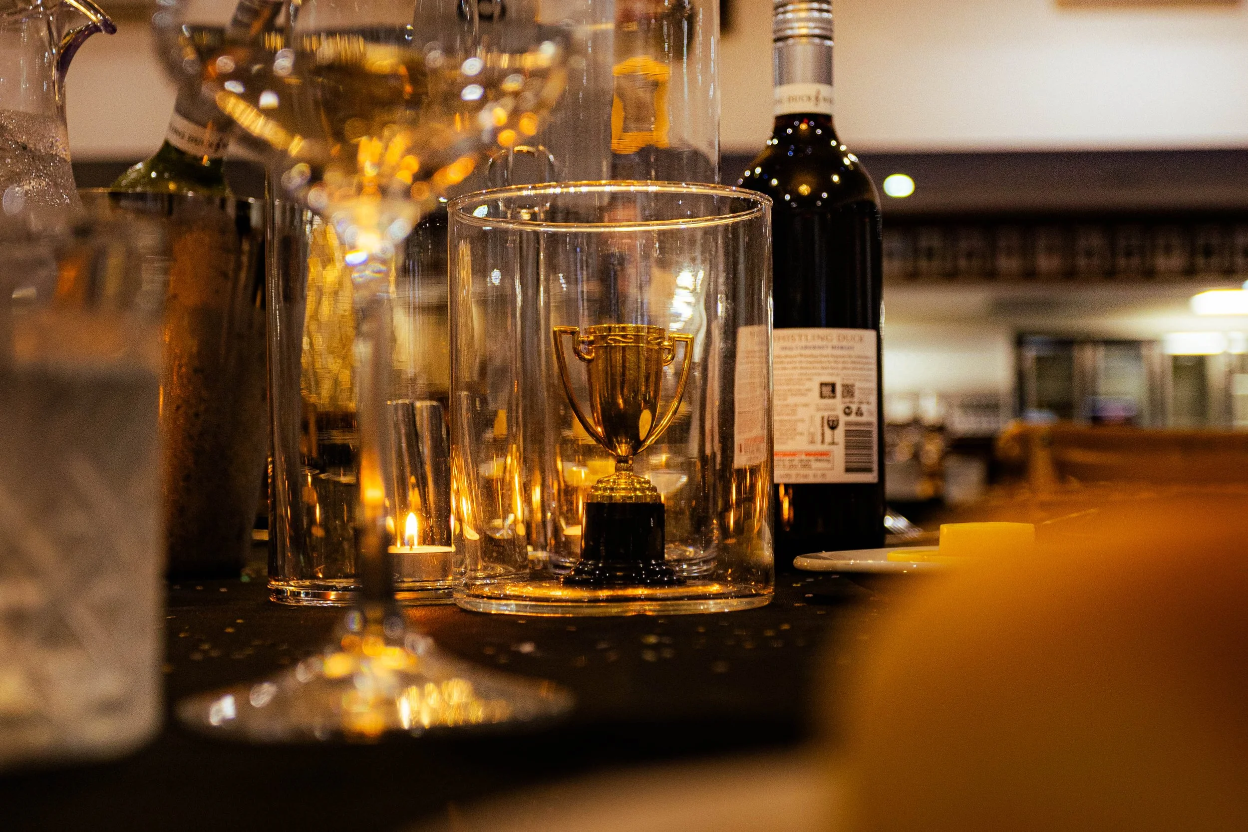 Close-up of a bar or restaurant counter with a small golden trophy figurine inside a glass, surrounded by bottles and drinkware, with warm lighting creating reflections and bokeh effects.