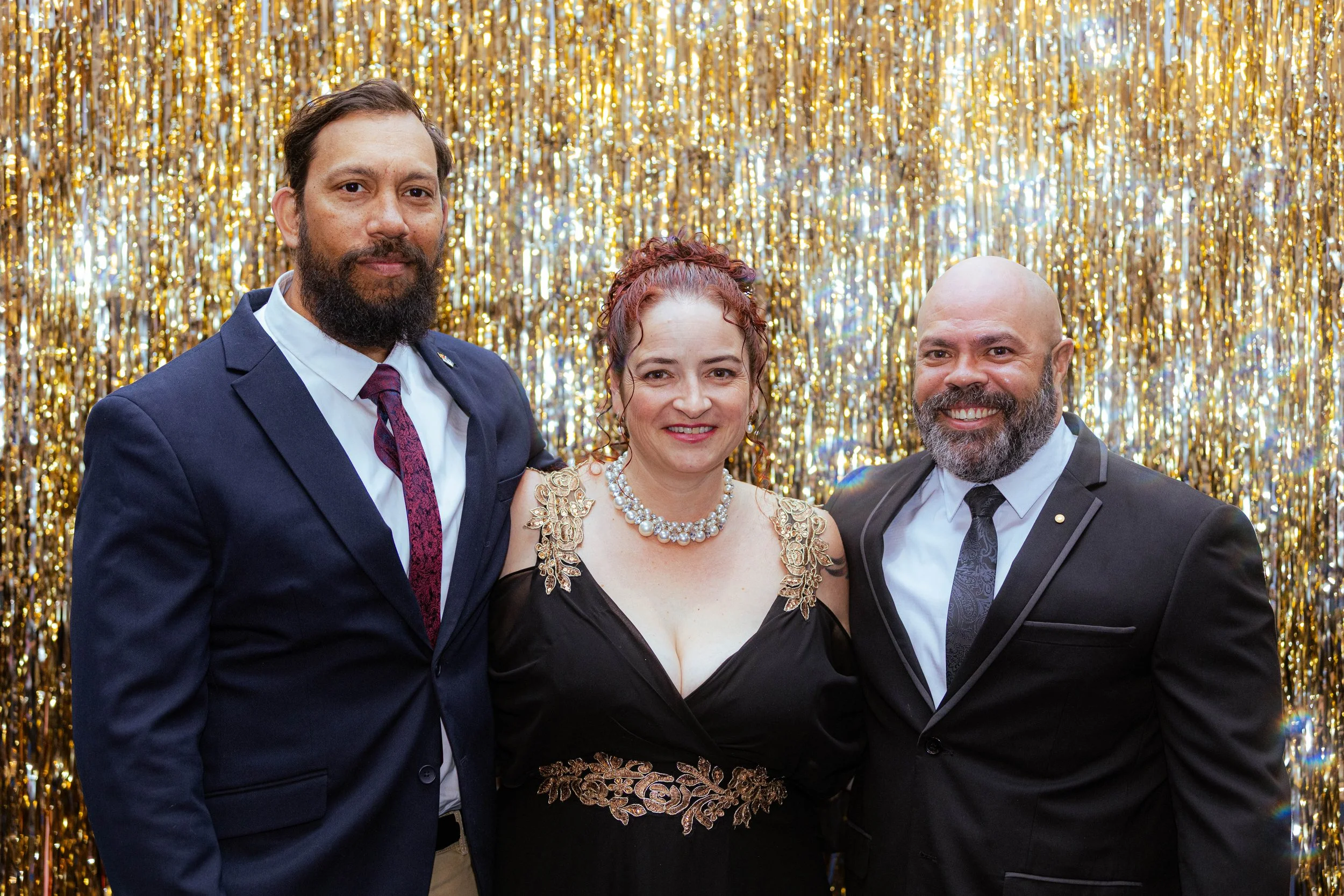 Three people in formal attire standing in front of a gold tinsel backdrop, smiling at the camera. The person in the middle is a woman wearing a black dress with gold embroidery, pearl necklace, and colorful earrings. The men on either side are both w
