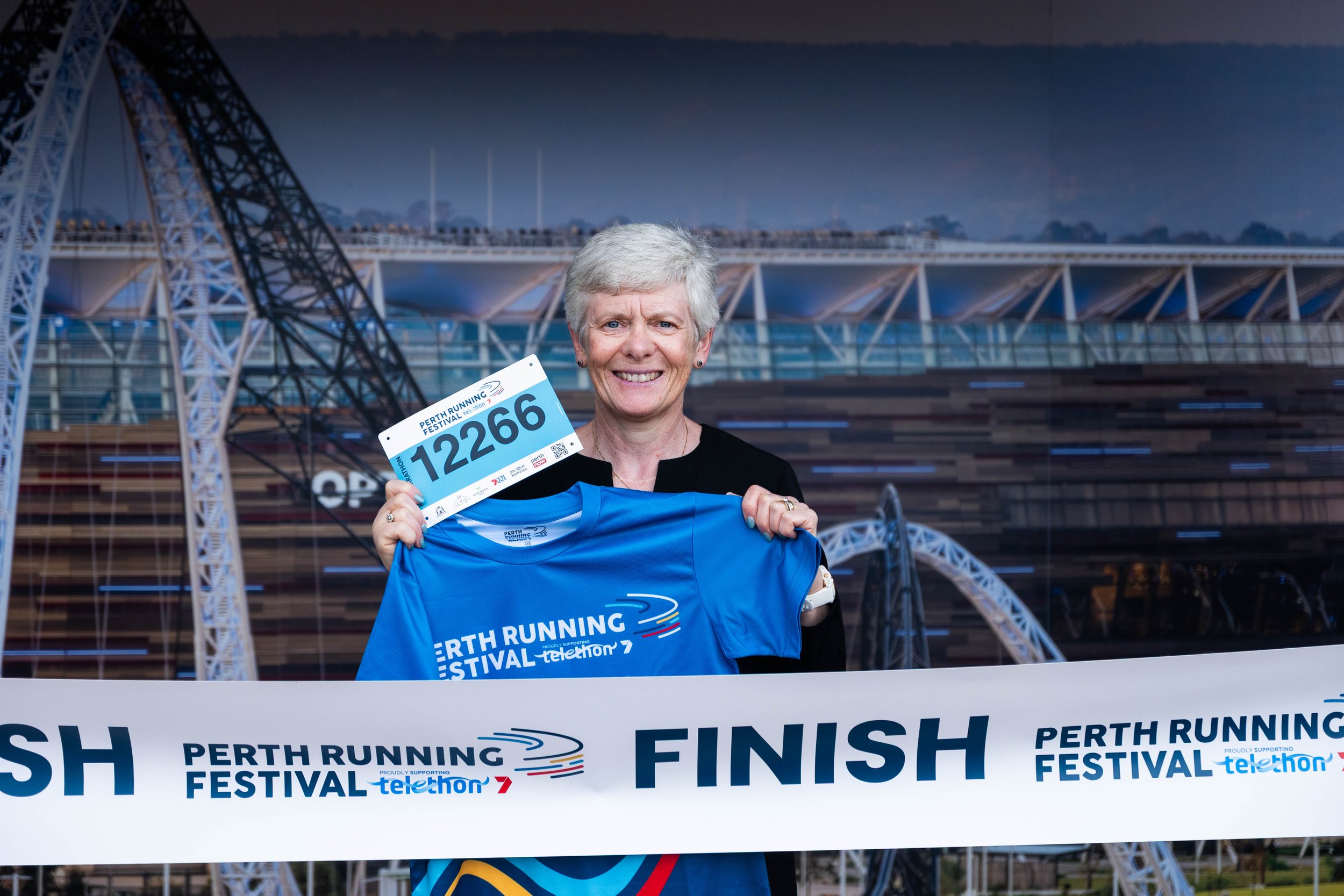 A smiling woman holding a race bib and a blue T-shirt stands behind the finish line at the Perth Running Festival, with a modern architectural structure in the background.