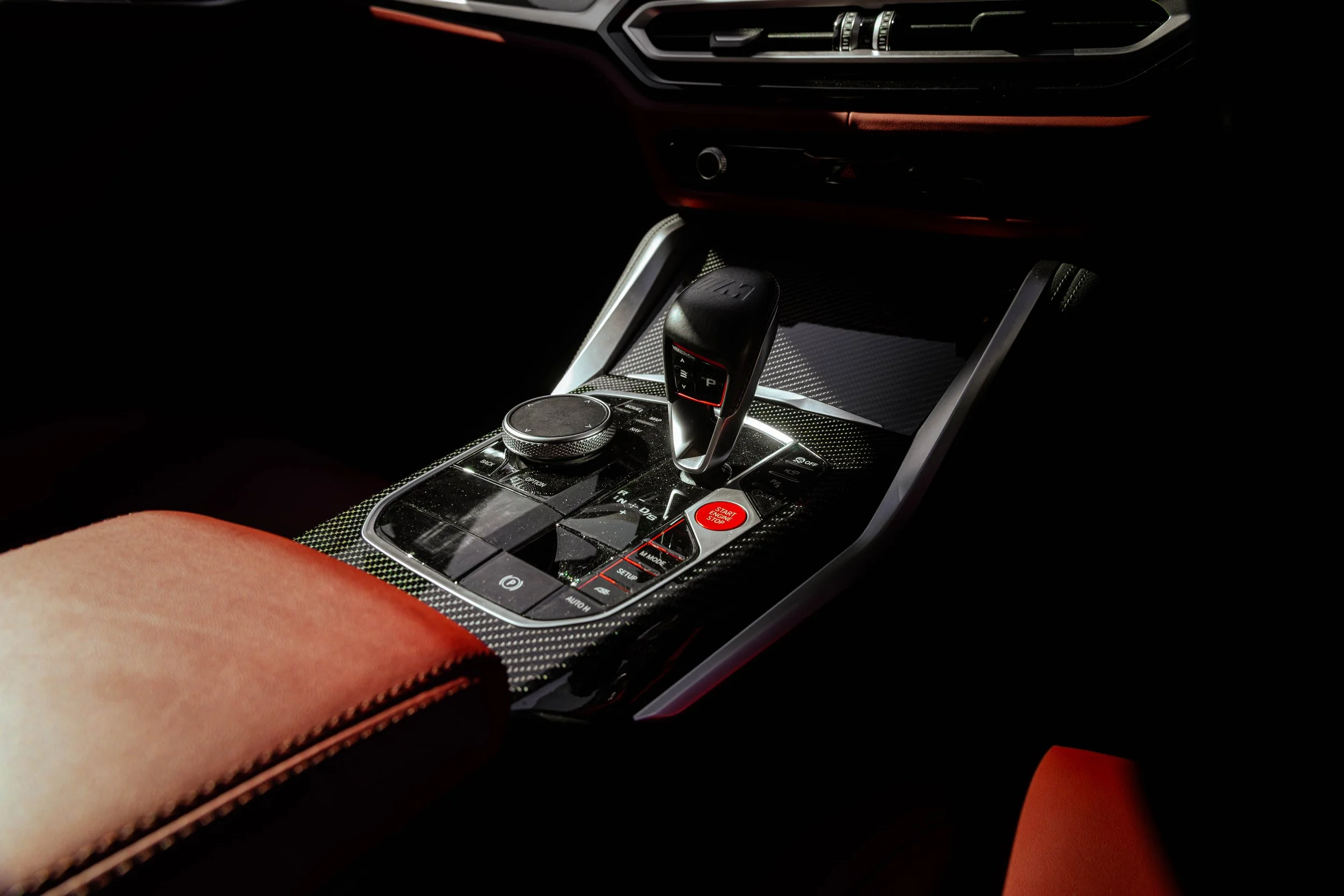 Close-up of a car's center console featuring a gear shifter, control buttons, and a large round control knob, with red seating visible in the foreground.