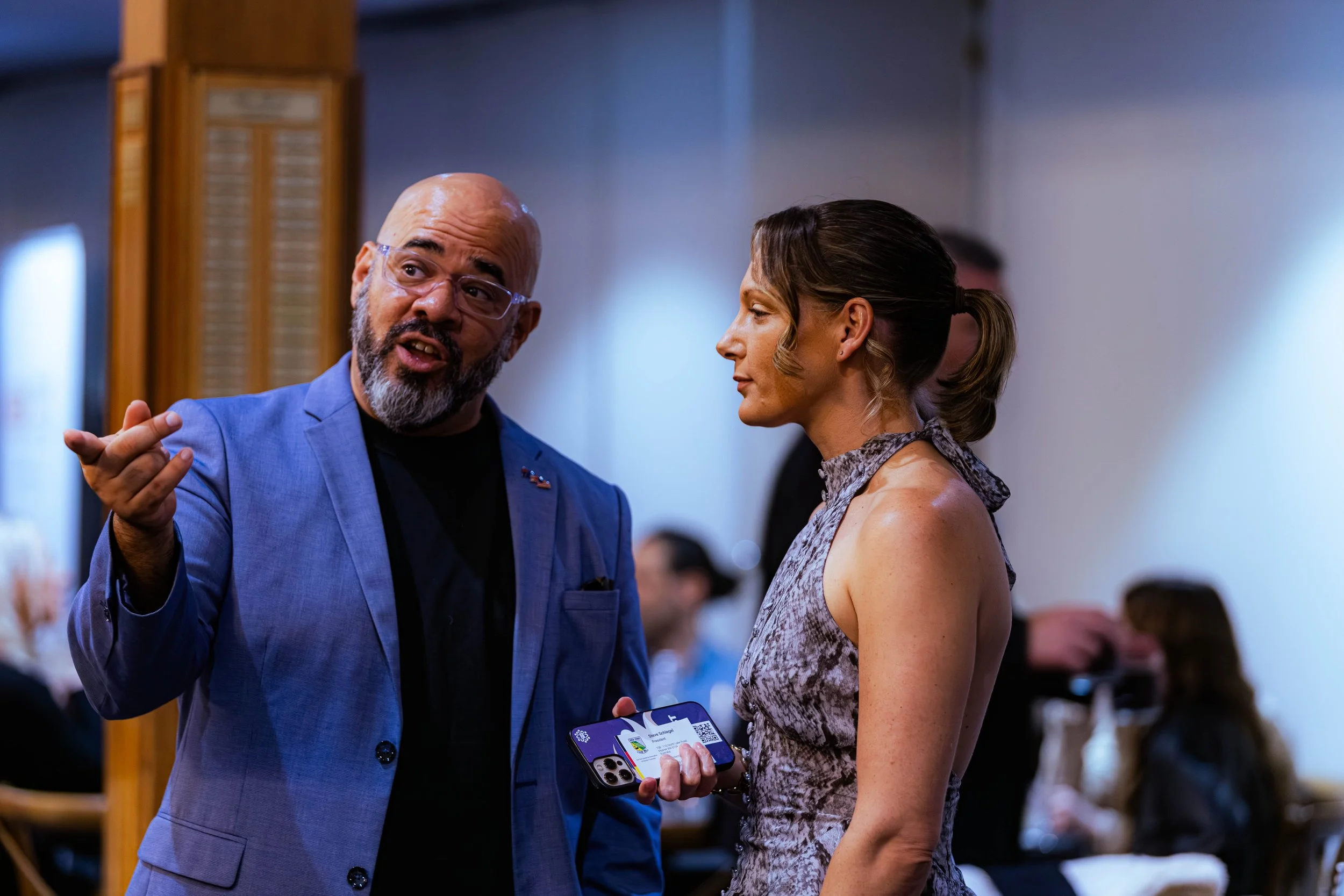 A man with glasses and a beard in a blue suit is talking to a woman with styled hair in a sleeveless dress at an indoor event. The woman is holding a phone with a QR code on it, and both are engaged in conversation.