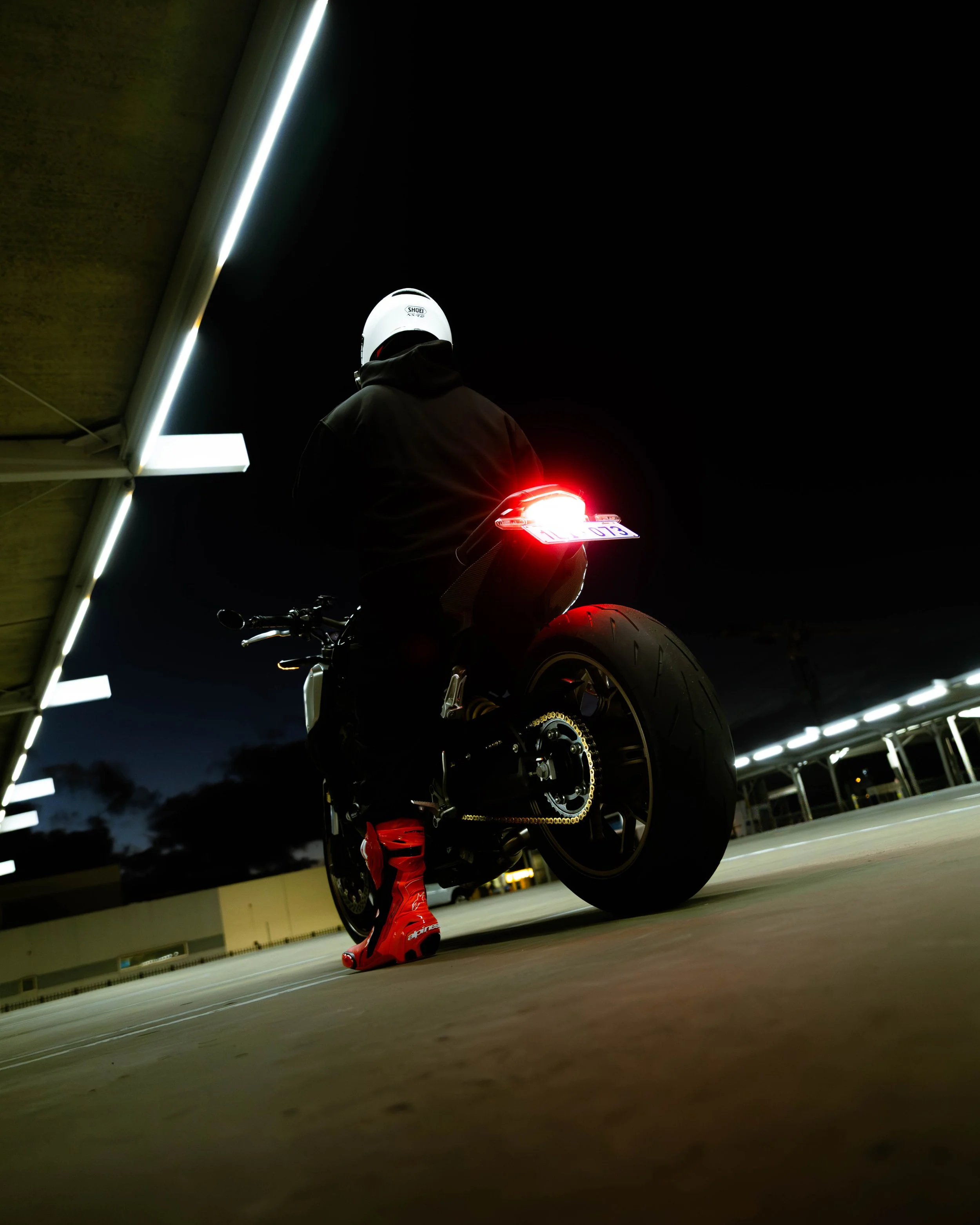 A person wearing a racing suit and red racing shoes, standing next to a motorcycle under a parking structure at night, with their back facing the camera, and a motorcycle license plate visible.