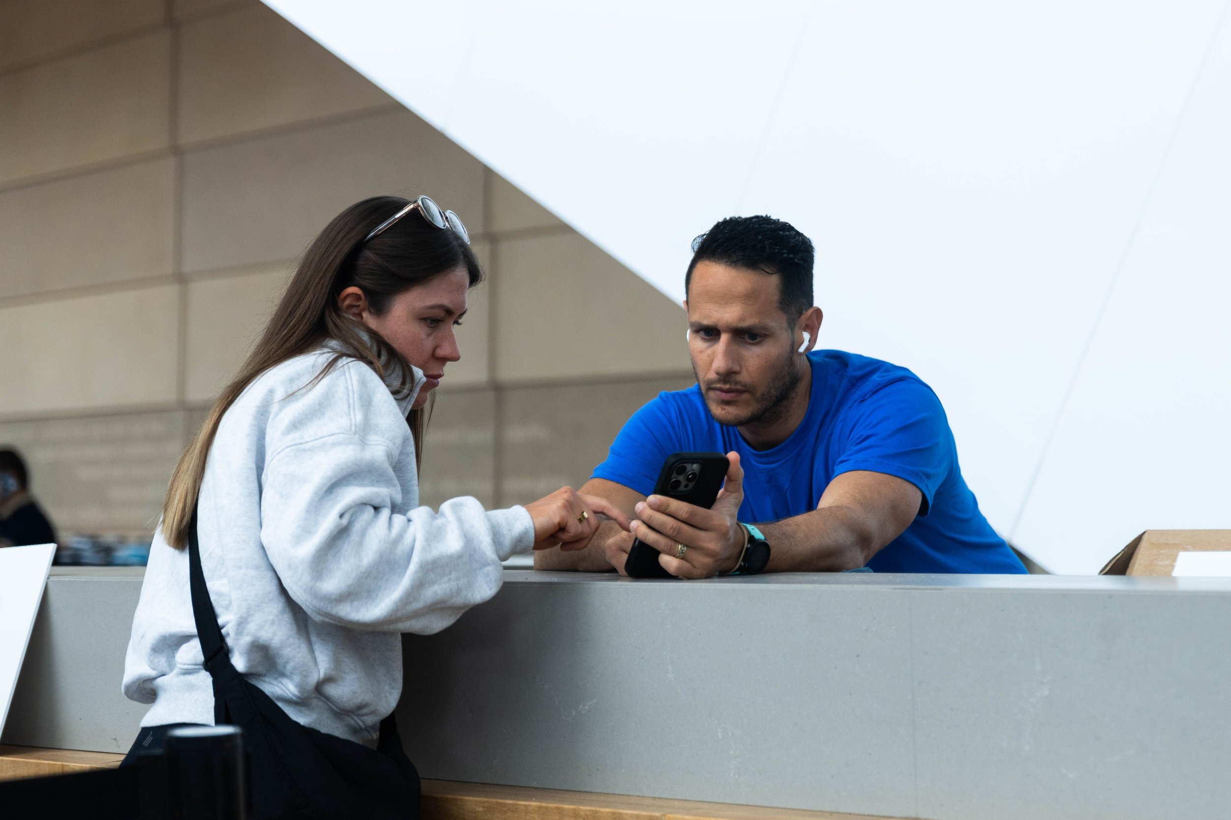 A woman and a man are sitting at a counter, looking at a smartphone together. The woman is wearing a gray hoodie and sunglasses on her head. The man is wearing a blue shirt and wireless earbuds. They are in an indoor setting with a modern ceiling des