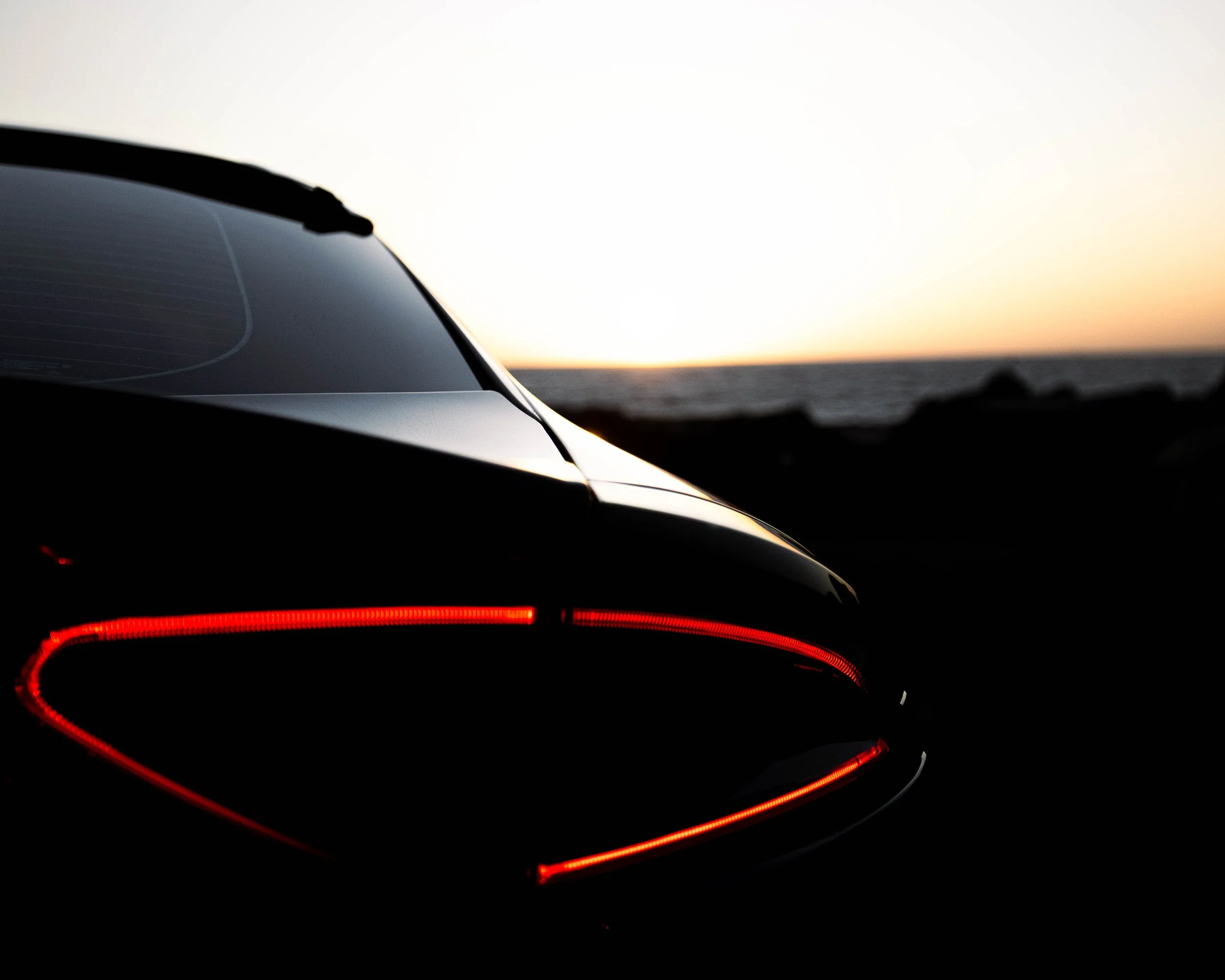 Rear view of a sports car with illuminated red tail lights against a sunset sky over the ocean.
