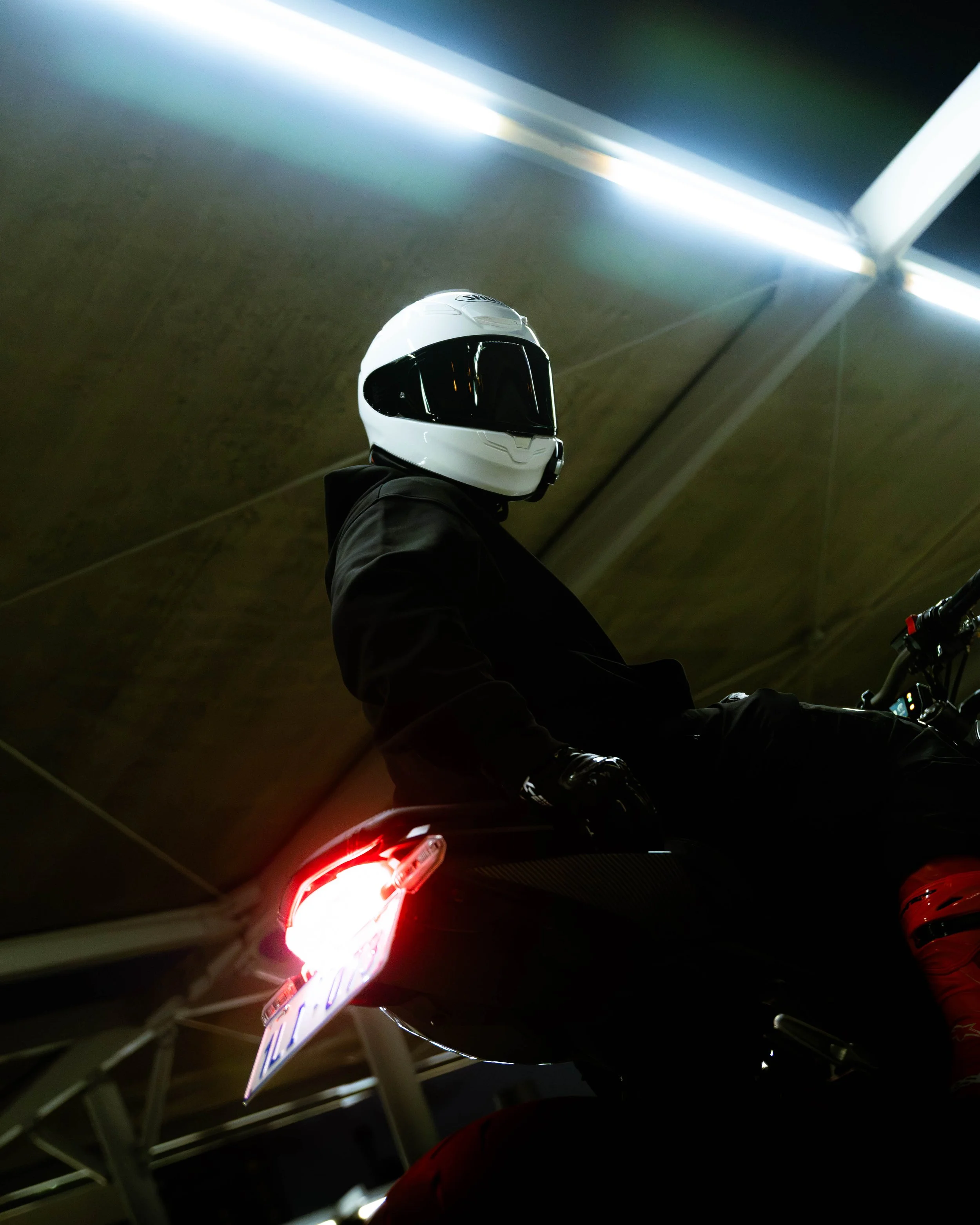 View of a person riding a motorcycle in a tunnel, wearing a white helmet with a black visor, illuminated by overhead lights, with the motorcycle's tail light glowing red.