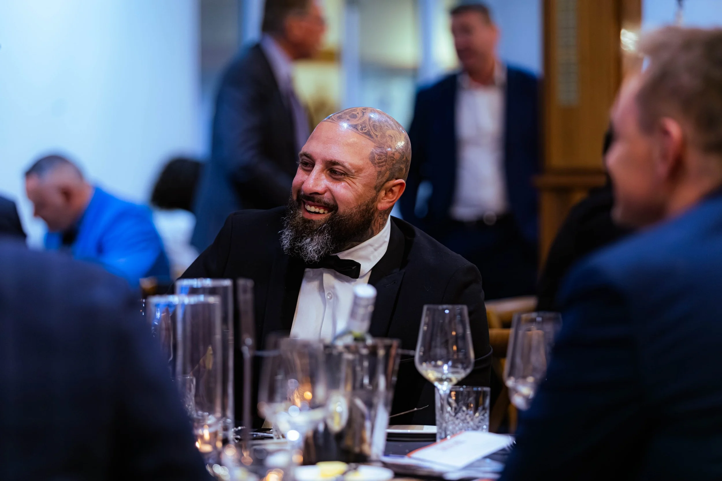 Smiling man with tattoos and beard dressed in a tuxedo at a formal event, with other guests in the background, seated at a dinner table with glasses and plates.