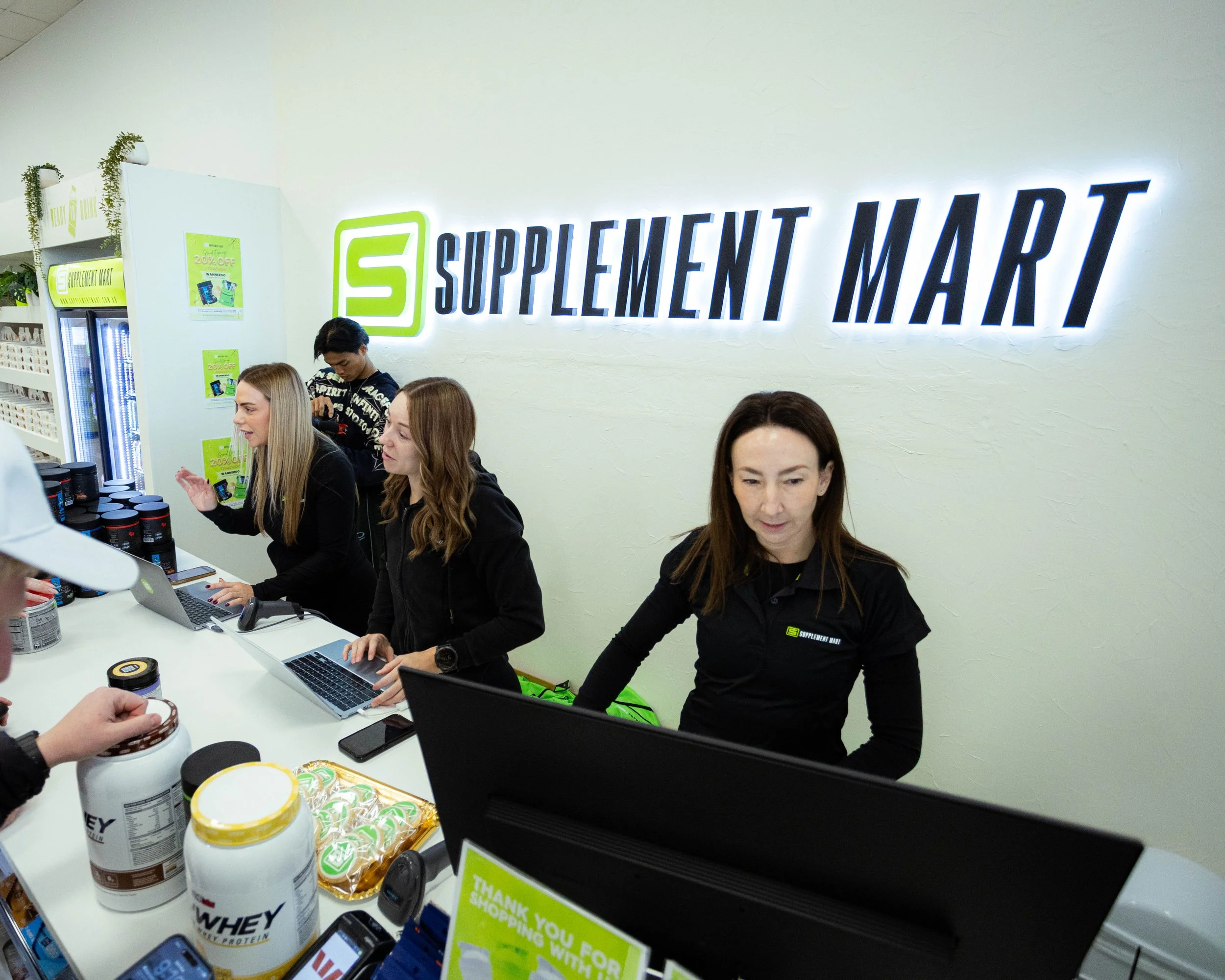 Interior of a supplement market with people working behind a counter, shelves with supplements and a large illuminated sign that reads 'Supplement Market'.
