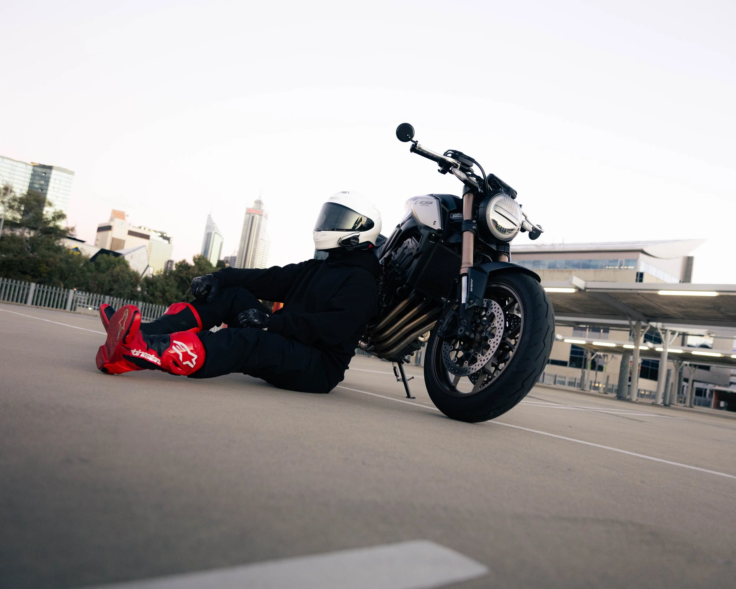 A person sitting on the ground next to a black motorcycle with a white helmet resting on the motorcycle handlebar. The person is wearing red shoes, black pants, and a black jacket. The background shows a city skyline with tall buildings and a parking