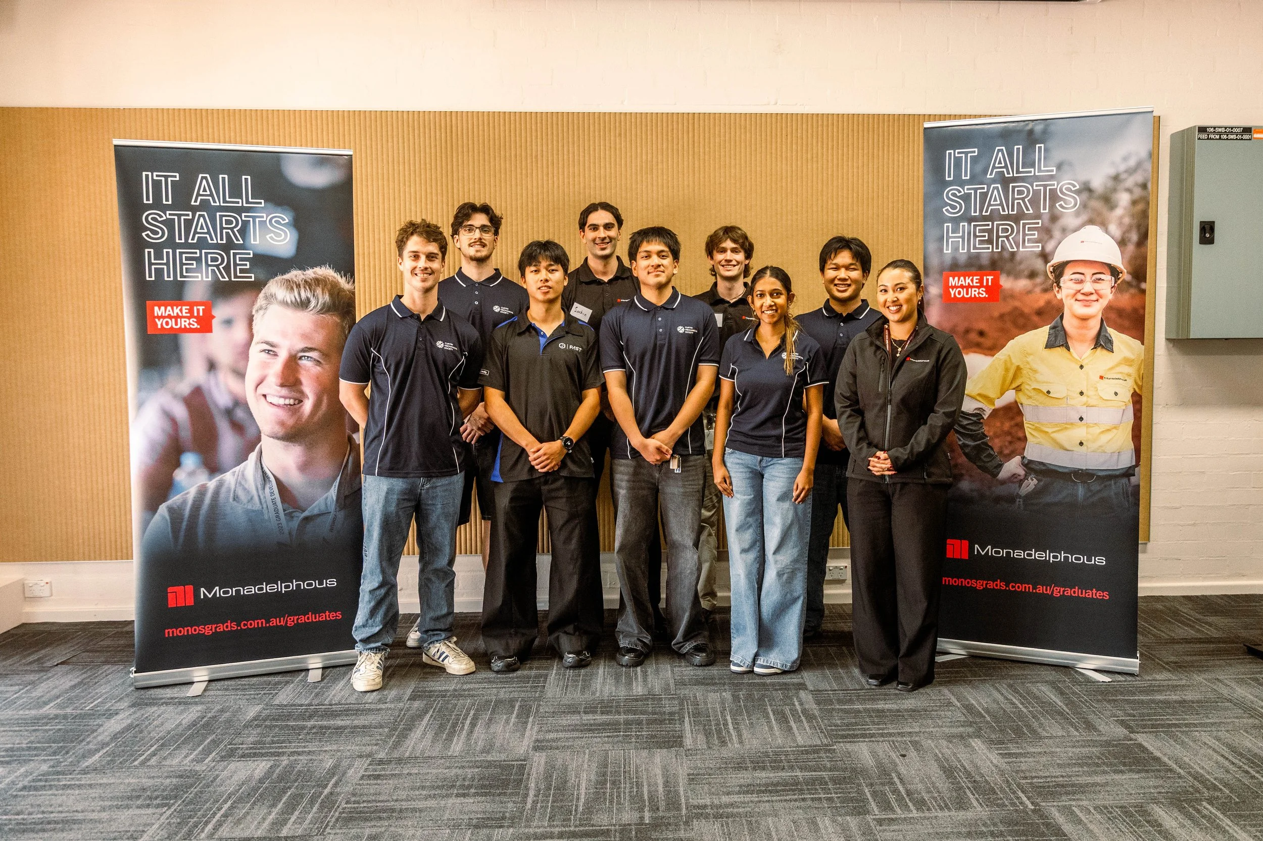 Group of ten young adults standing indoors in front of two large display banners, smiling at the camera. The banners display the phrases 'IT ALL STARTS HERE' and 'MAKE IT YOURS', with branding for Monadelphous and the web address monosgrads.com.au/gr