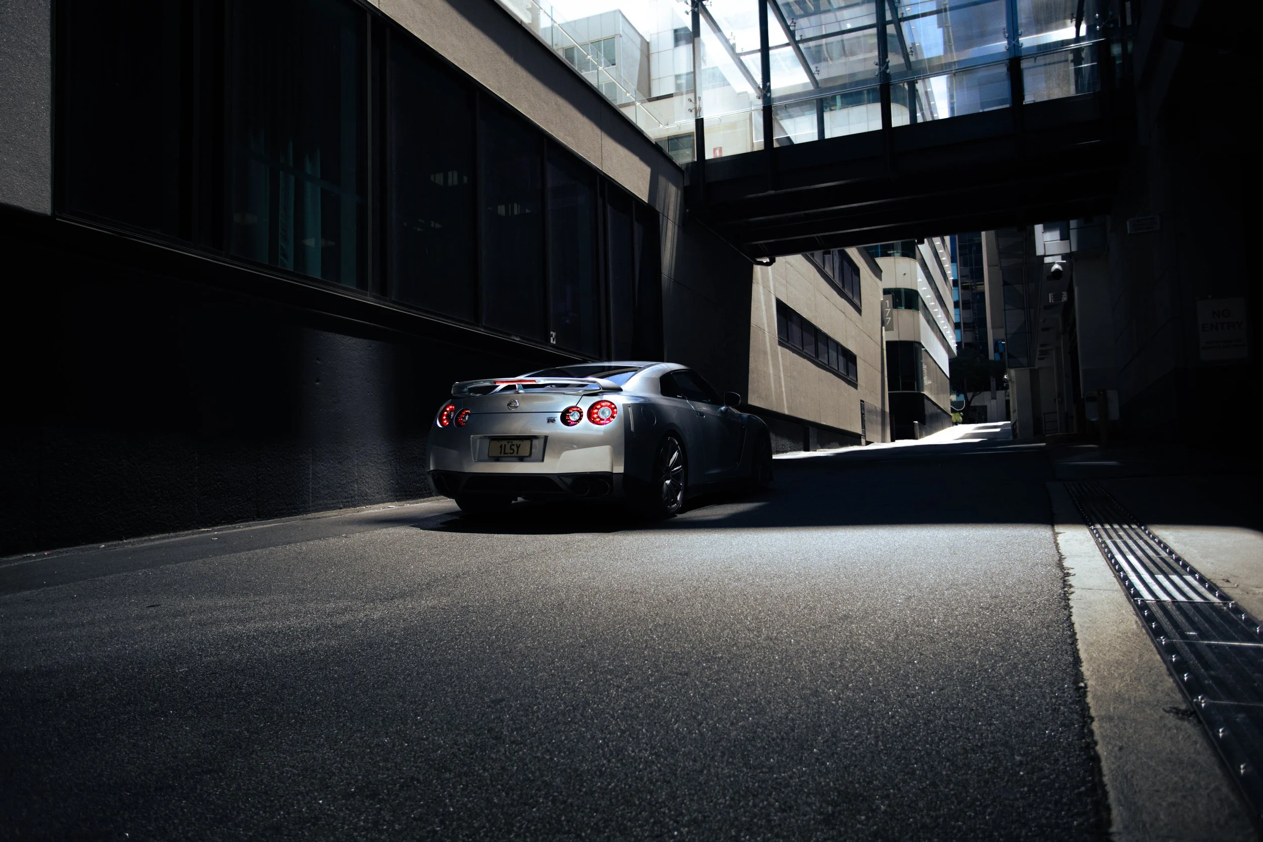 A silver sports car parked in a narrow urban alleyway surrounded by tall modern buildings with glass windows.