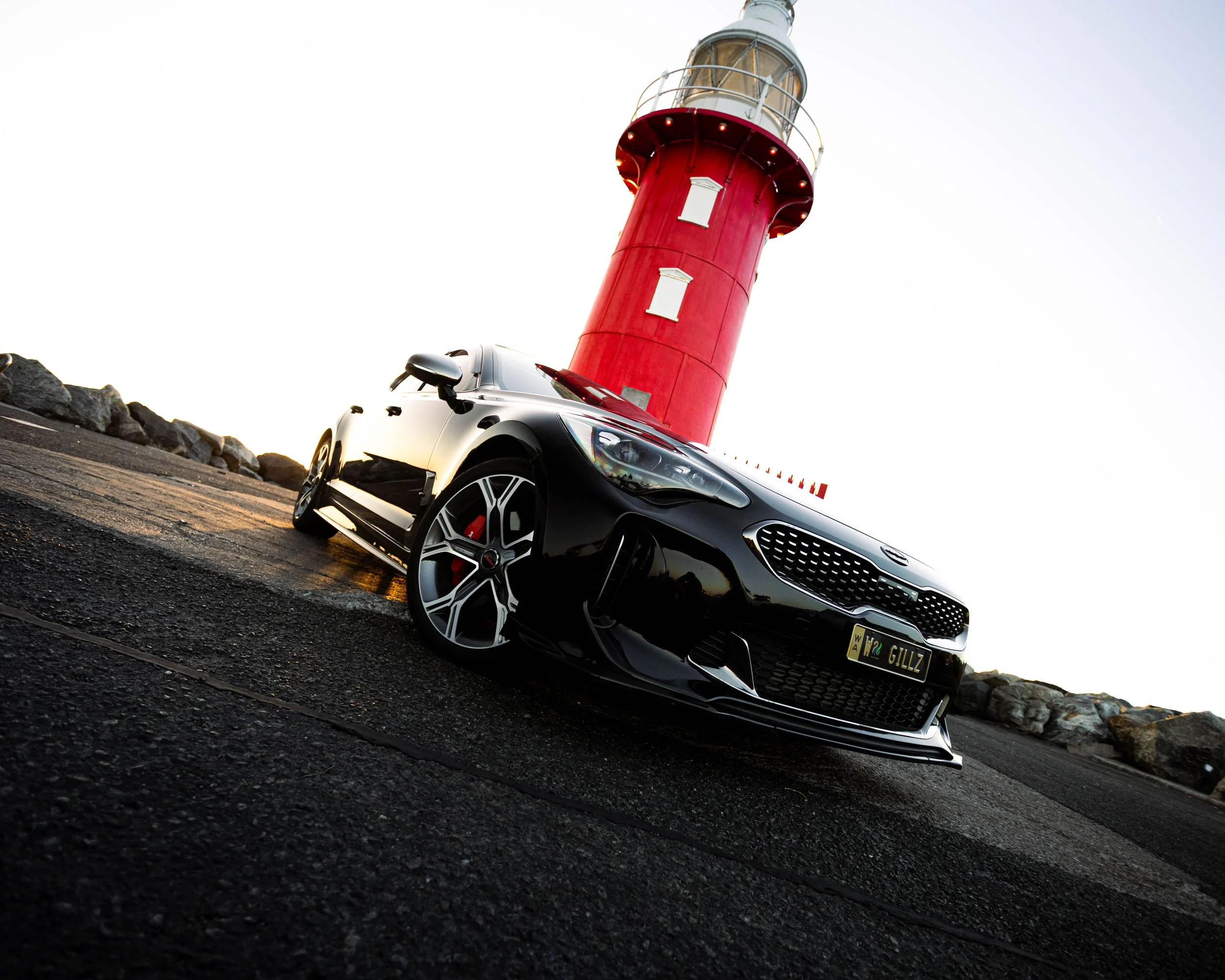 A black sports car parked beside a red lighthouse on a rocky shoreline during sunset.