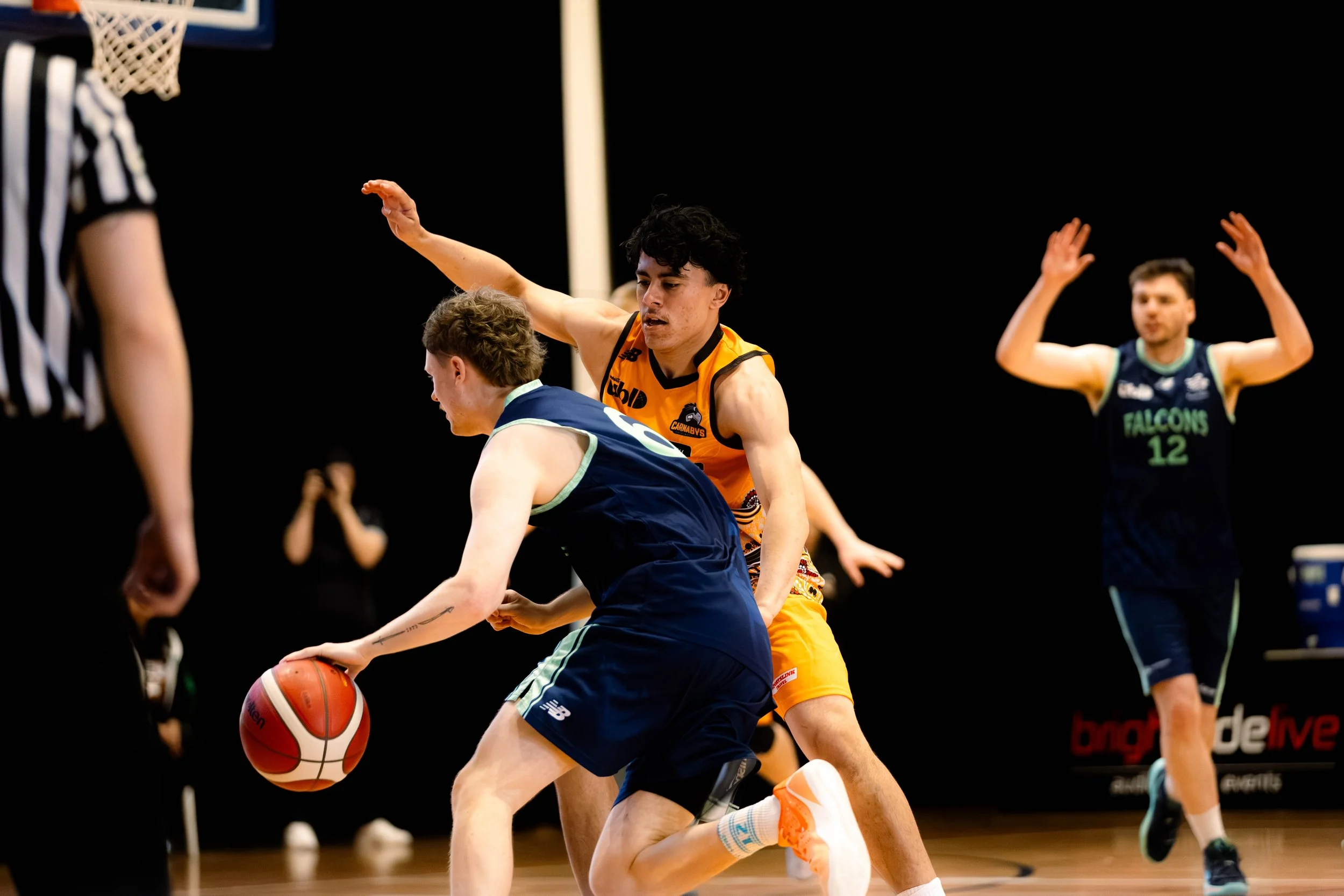 Two young male basketball players in action on the court, with one dribbling the ball and the other defending closely. An additional player and basketball hoop are visible in the background.