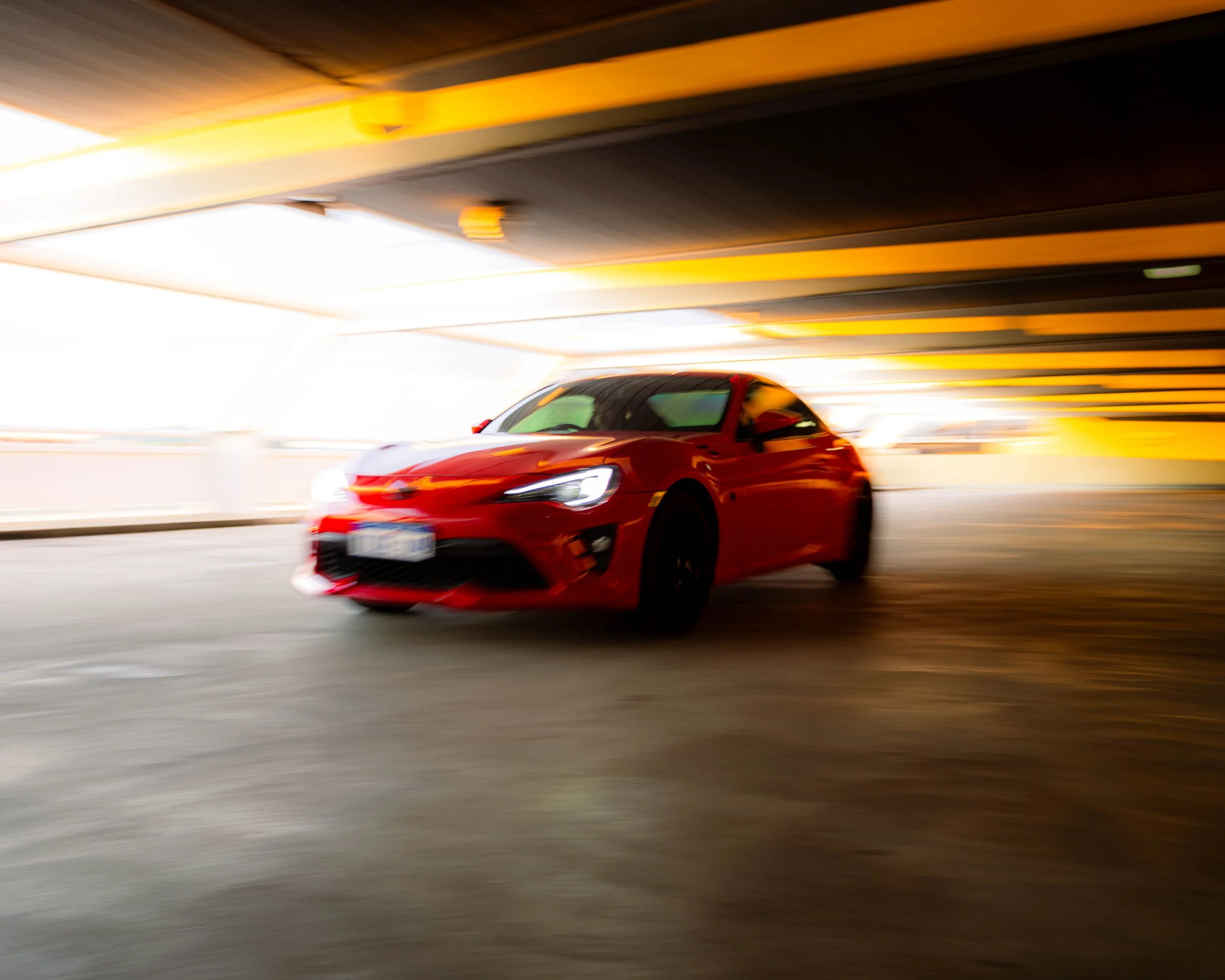 A red sports car driving through a brightly lit parking garage, with yellow and black painted ceiling beams visible above.