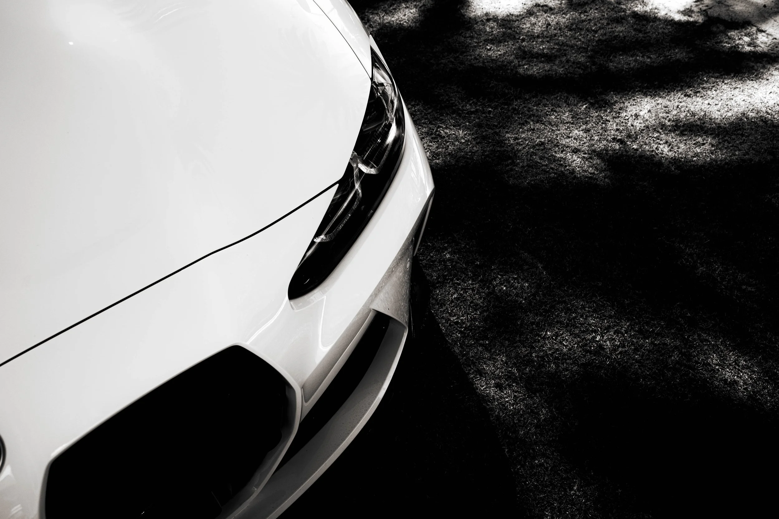 Close-up of the front of a white car parked on a rough-textured dark ground, with the car's headlight and part of the hood visible.