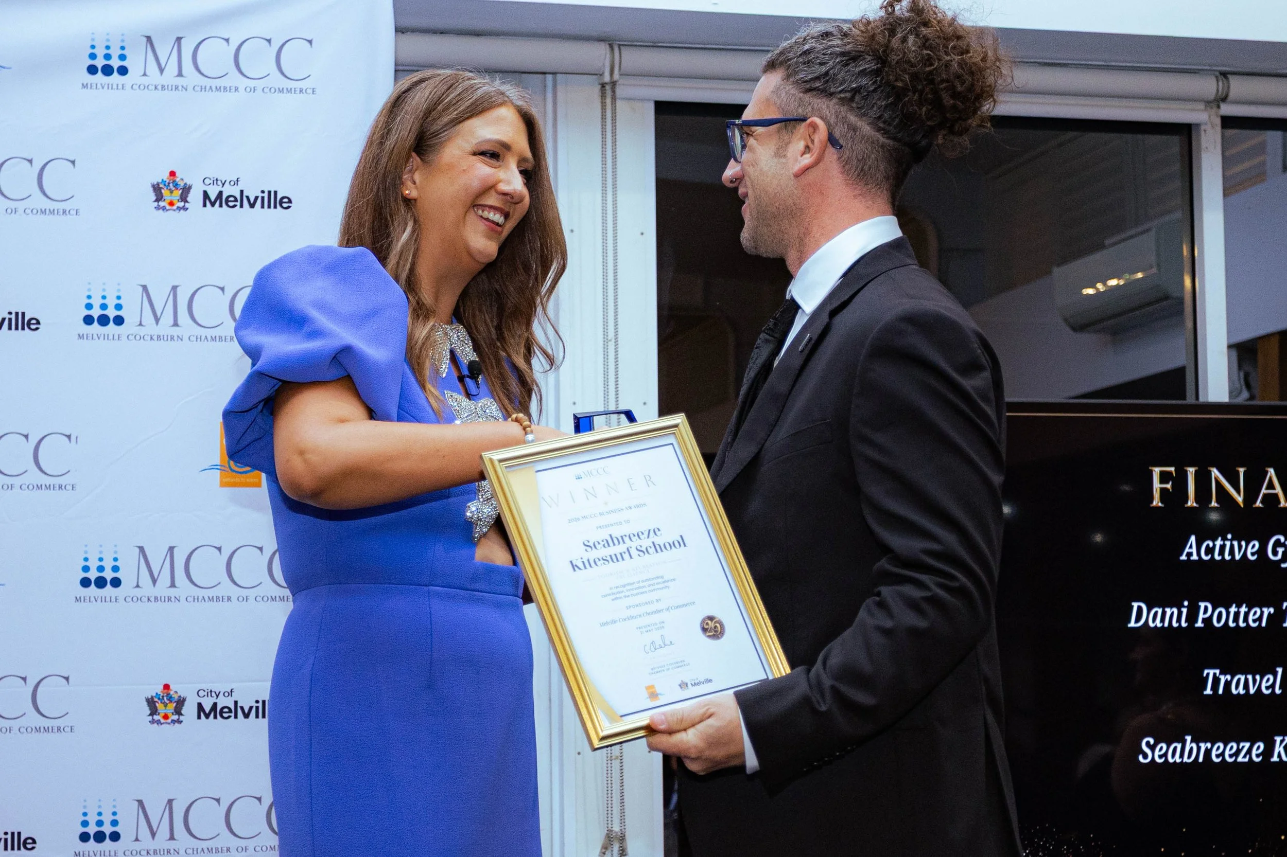 A woman in a blue dress receives an award certificate from a man in a black suit and glasses during a ceremony at the Melville Cockburn Chamber of Commerce event.
