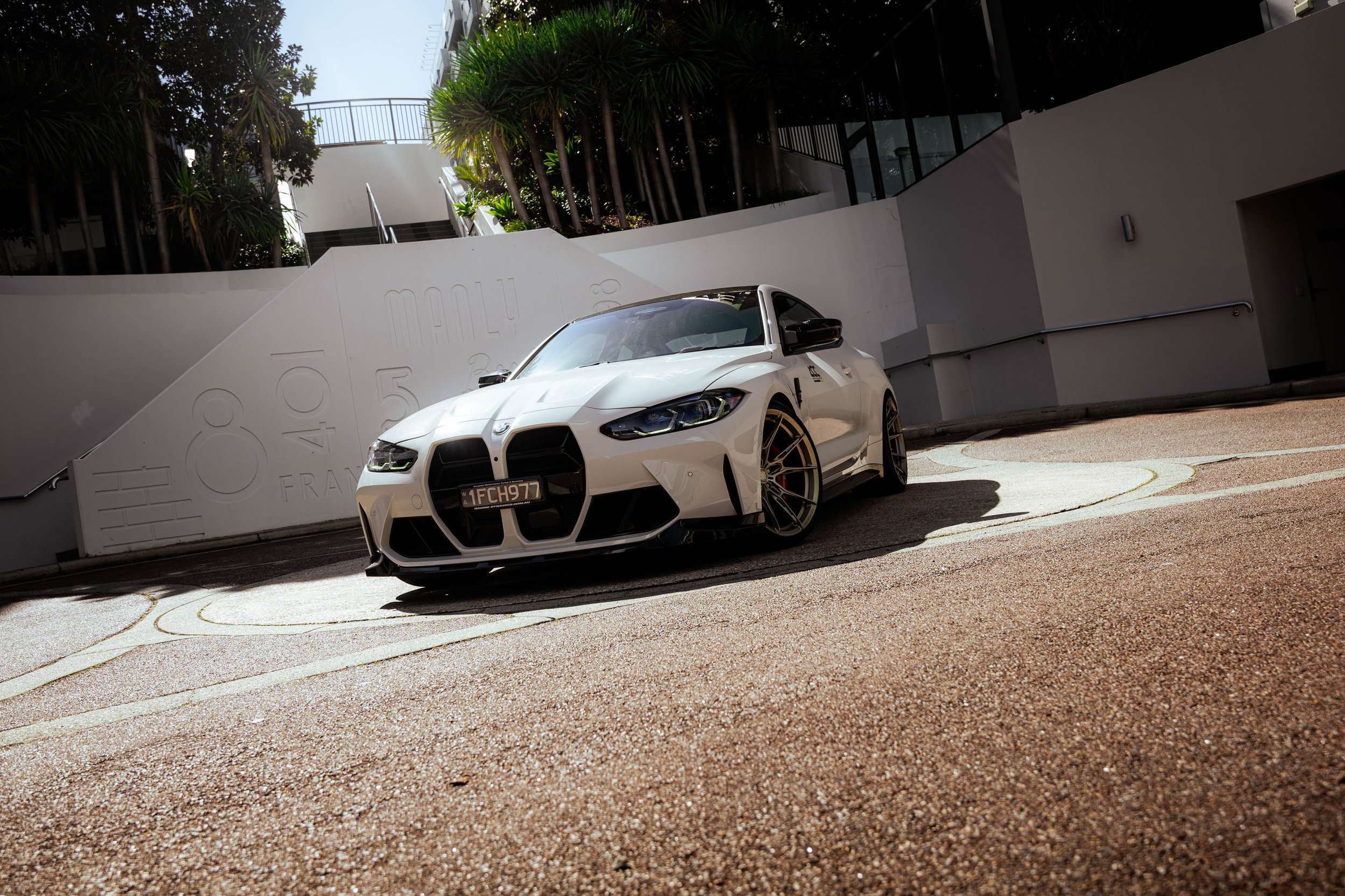 White sports car parked along a sidewalk in front of a modern building with trees and plants, and a white wall with embossed text.