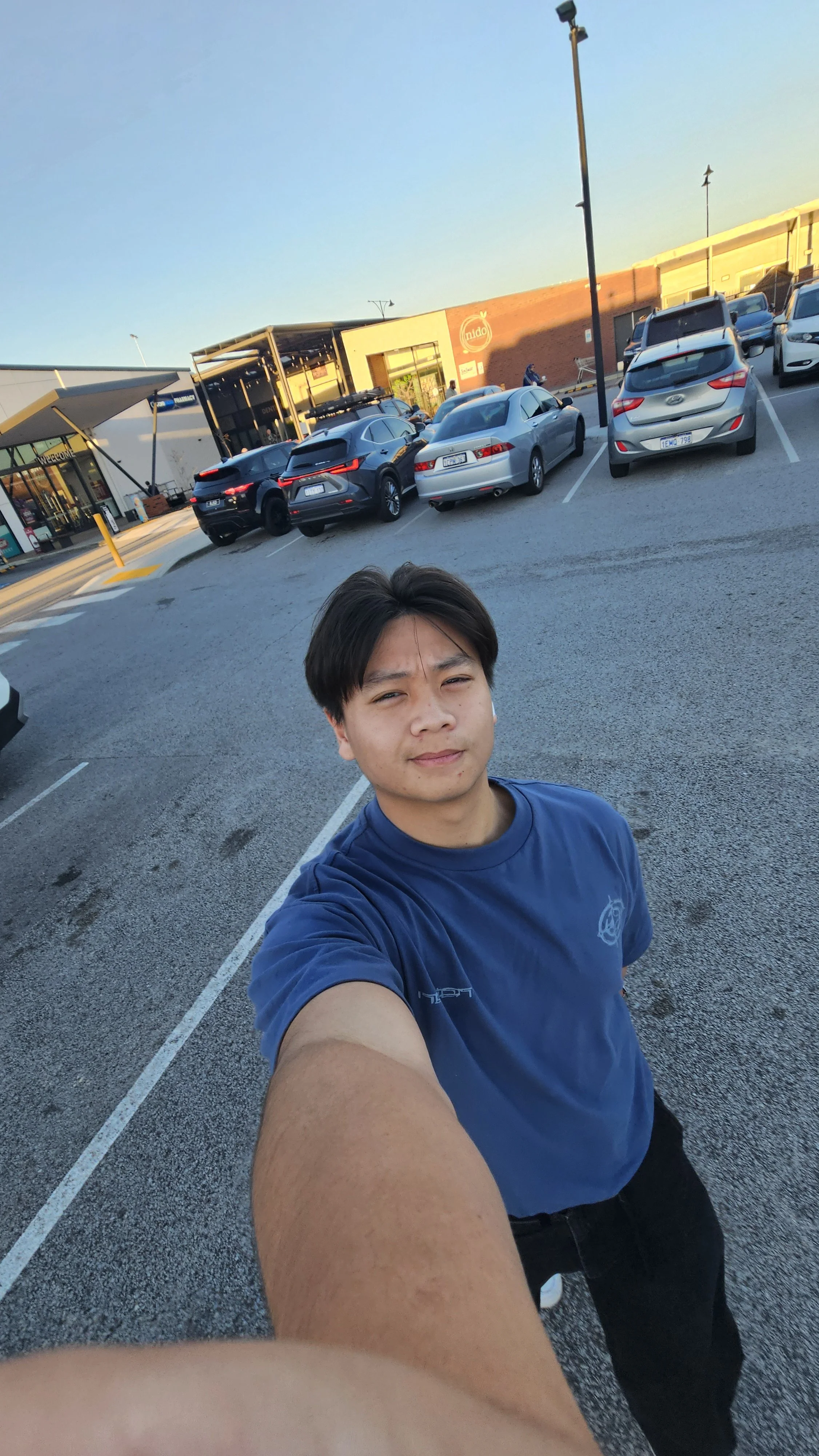 Selfie of a young man in a parking lot taking a photo with a smartphone. There are parked cars, a shopping center, and a clear sunset sky in the background.