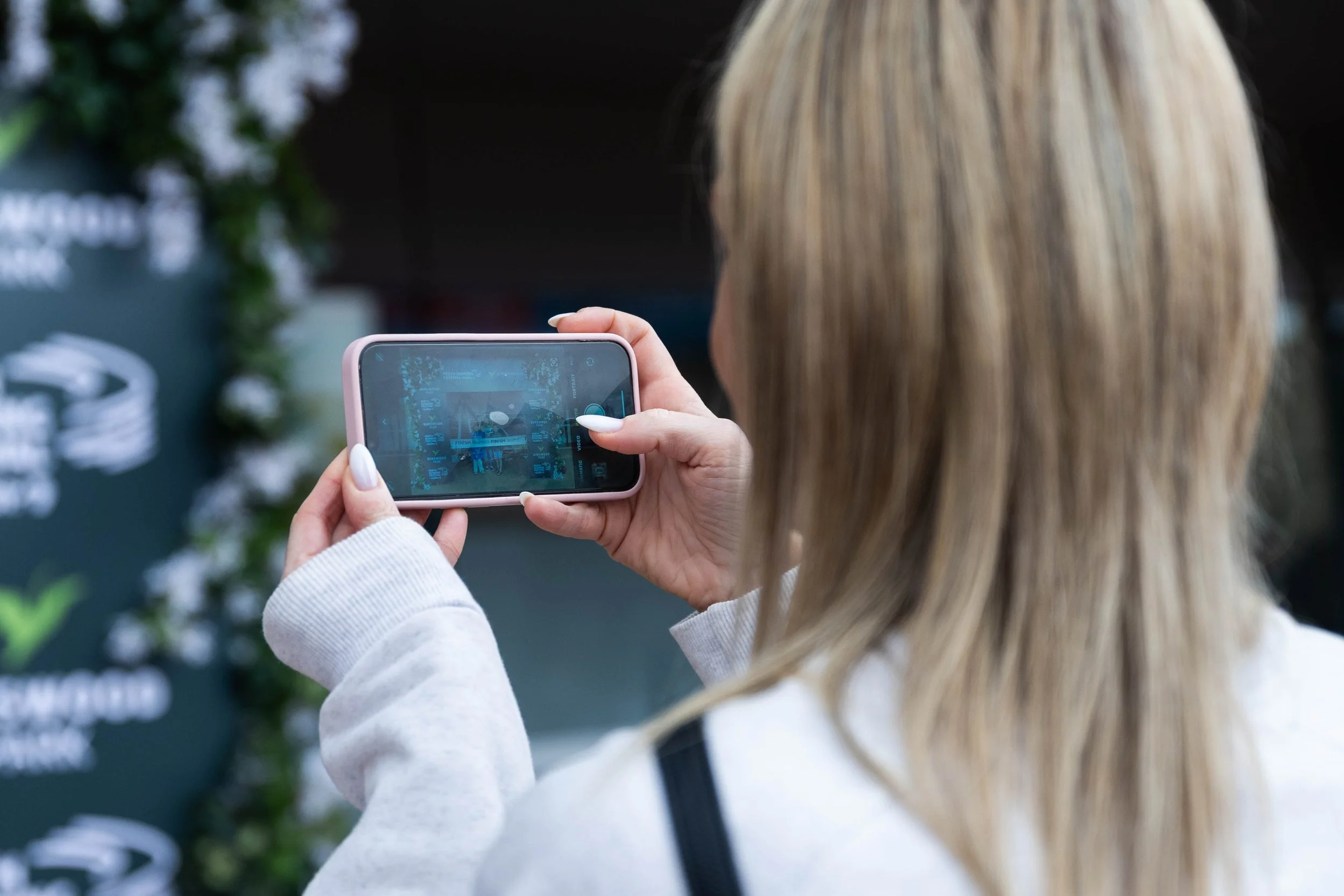 A person with blonde hair taking a photo or recording a video with a smartphone at an outdoor event.
