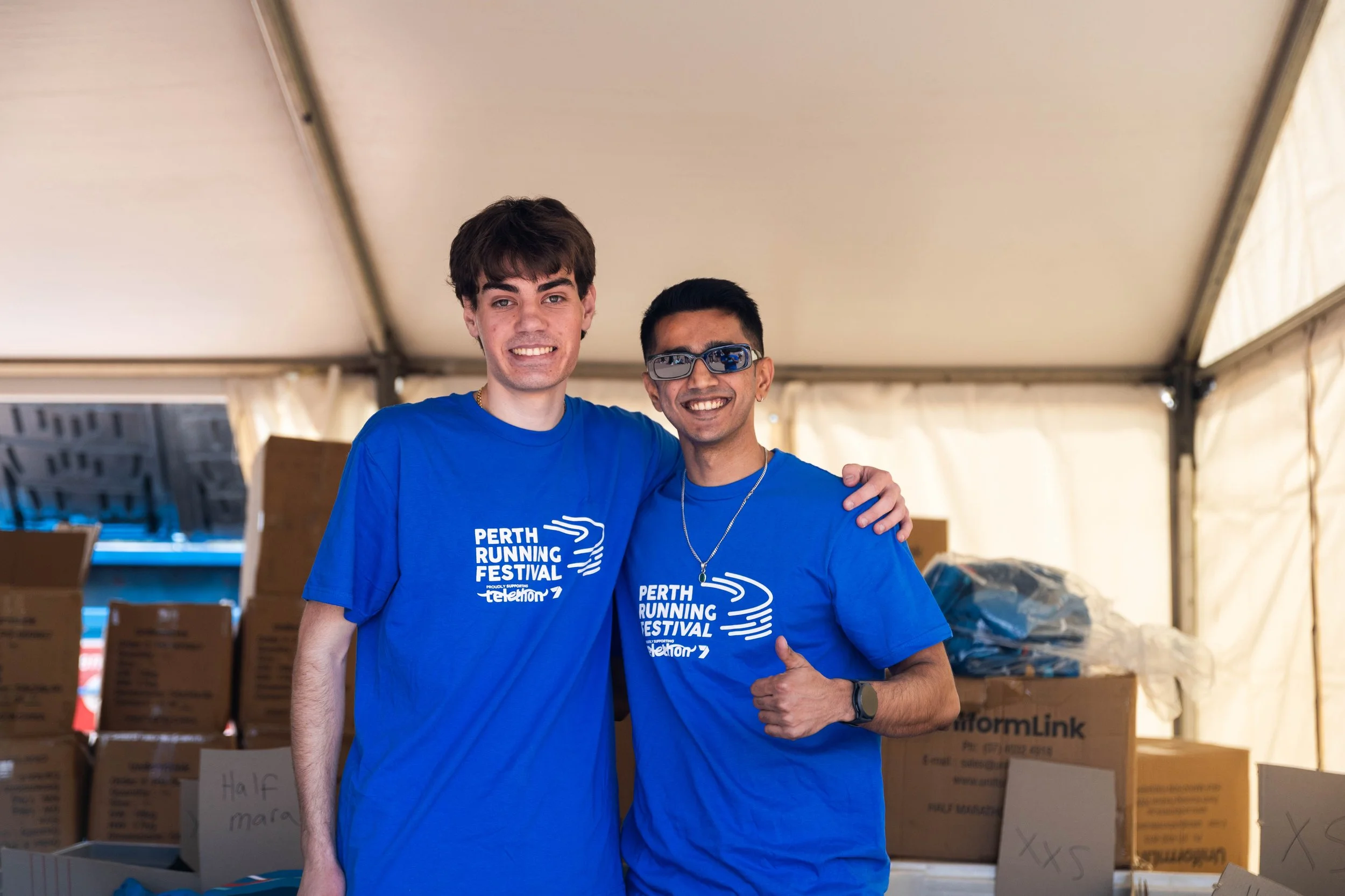 Two young men wearing blue Perth Running Festival T-shirts inside a tent, standing close together, smiling at the camera, with boxes in the background.