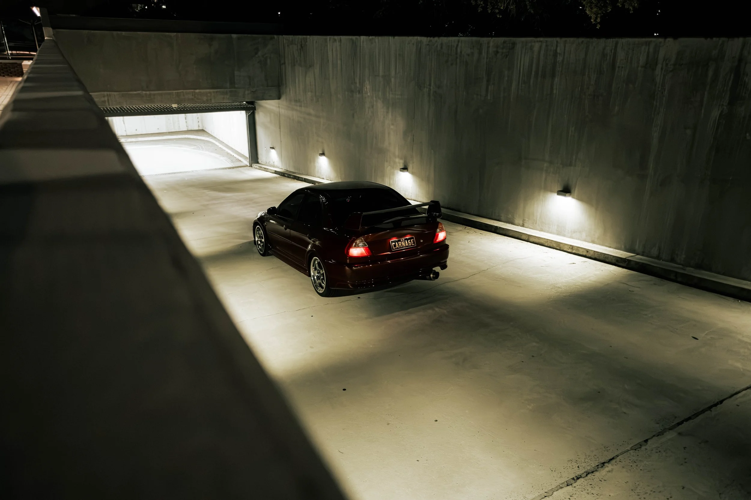 A black sports car parked in a dimly lit underground garage with concrete walls, illuminated by wall-mounted lights.