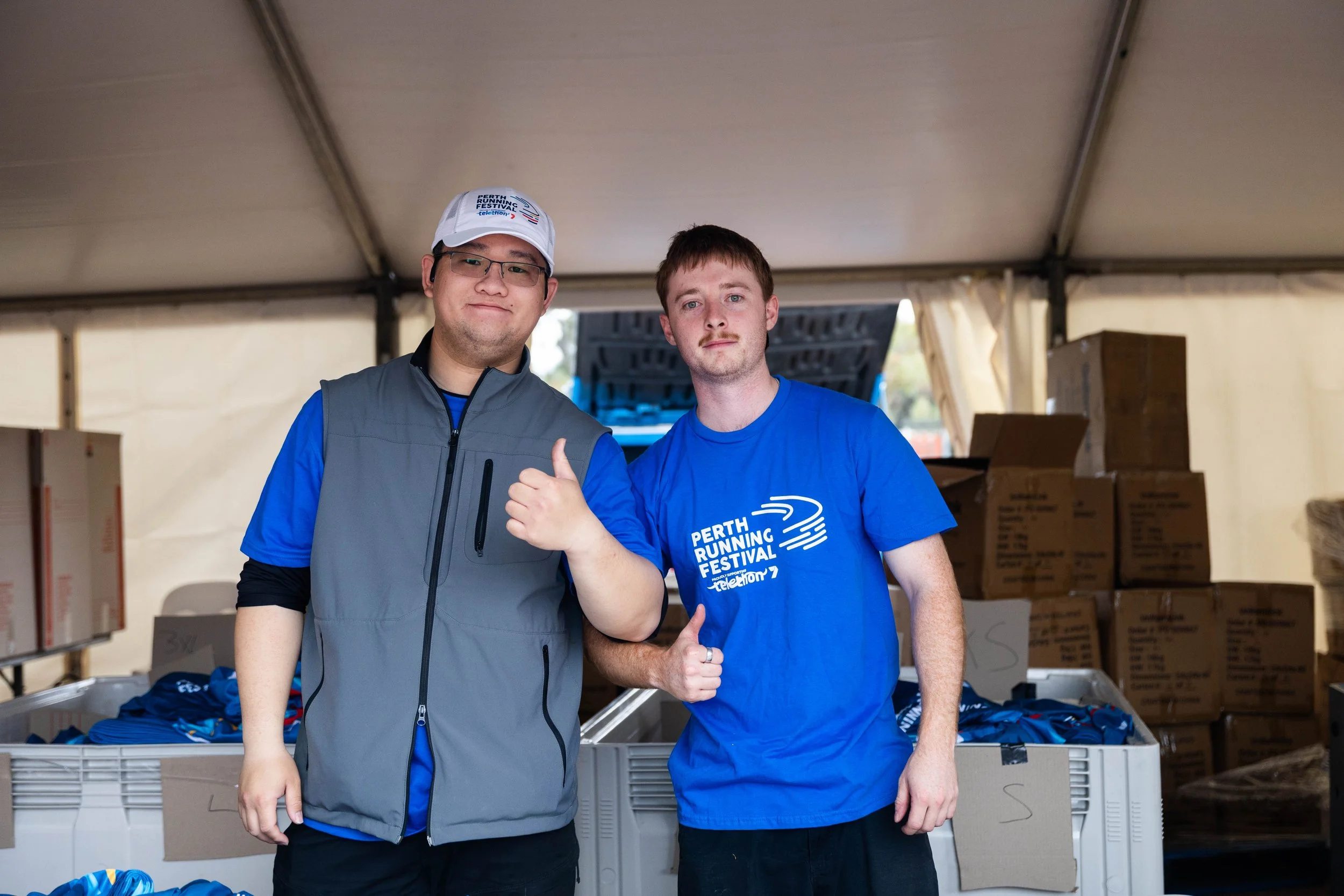 Two men standing inside a tent at an event, smiling at the camera. One is giving a thumbs-up gesture, and they are wearing T-shirts and a vest. The background has boxes and supplies, with some shirts visible on a table.