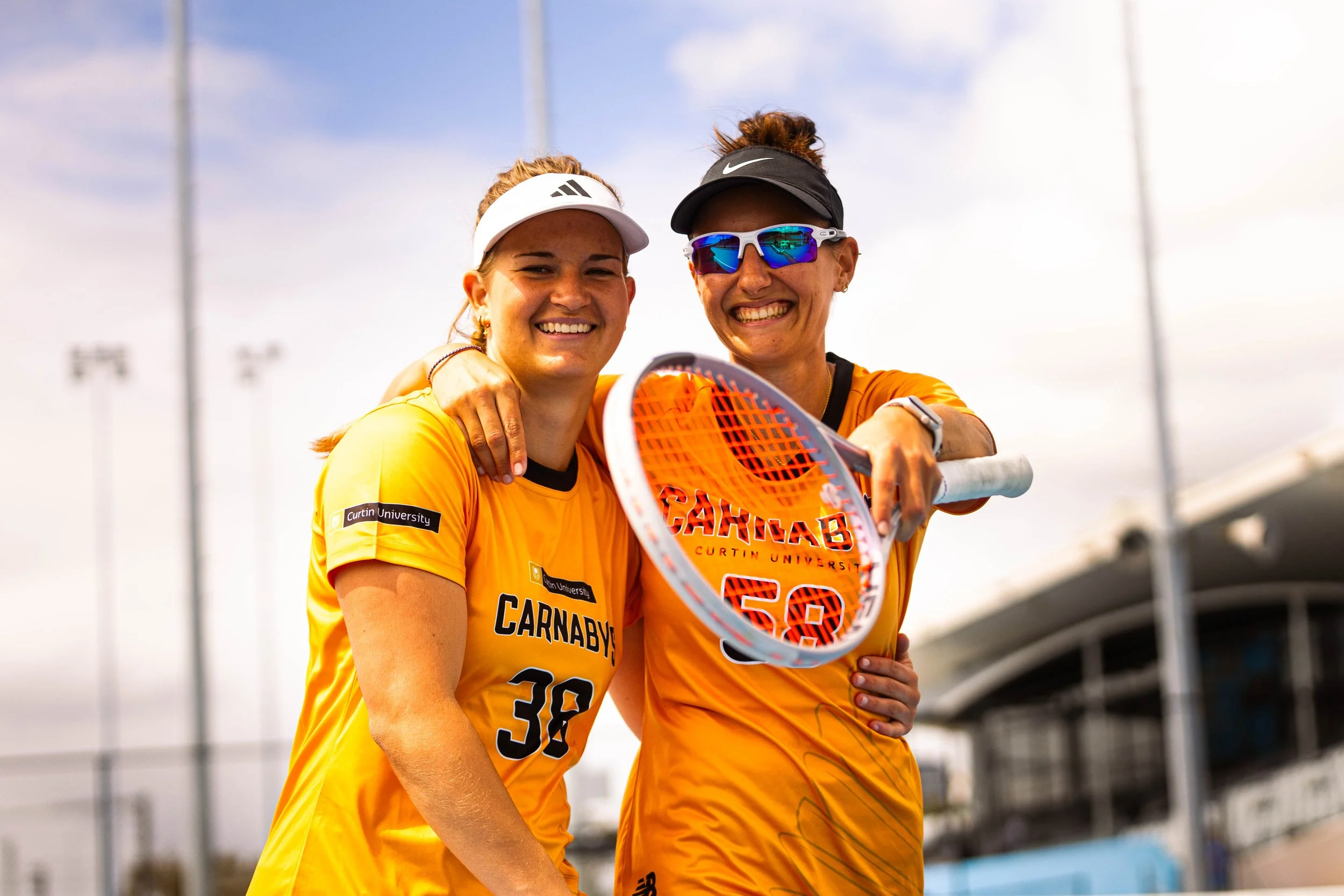 Two female athletes in yellow jerseys with 'Carnaby' on the front, smiling and posing with a tennis racket, outdoors on a sunny day.