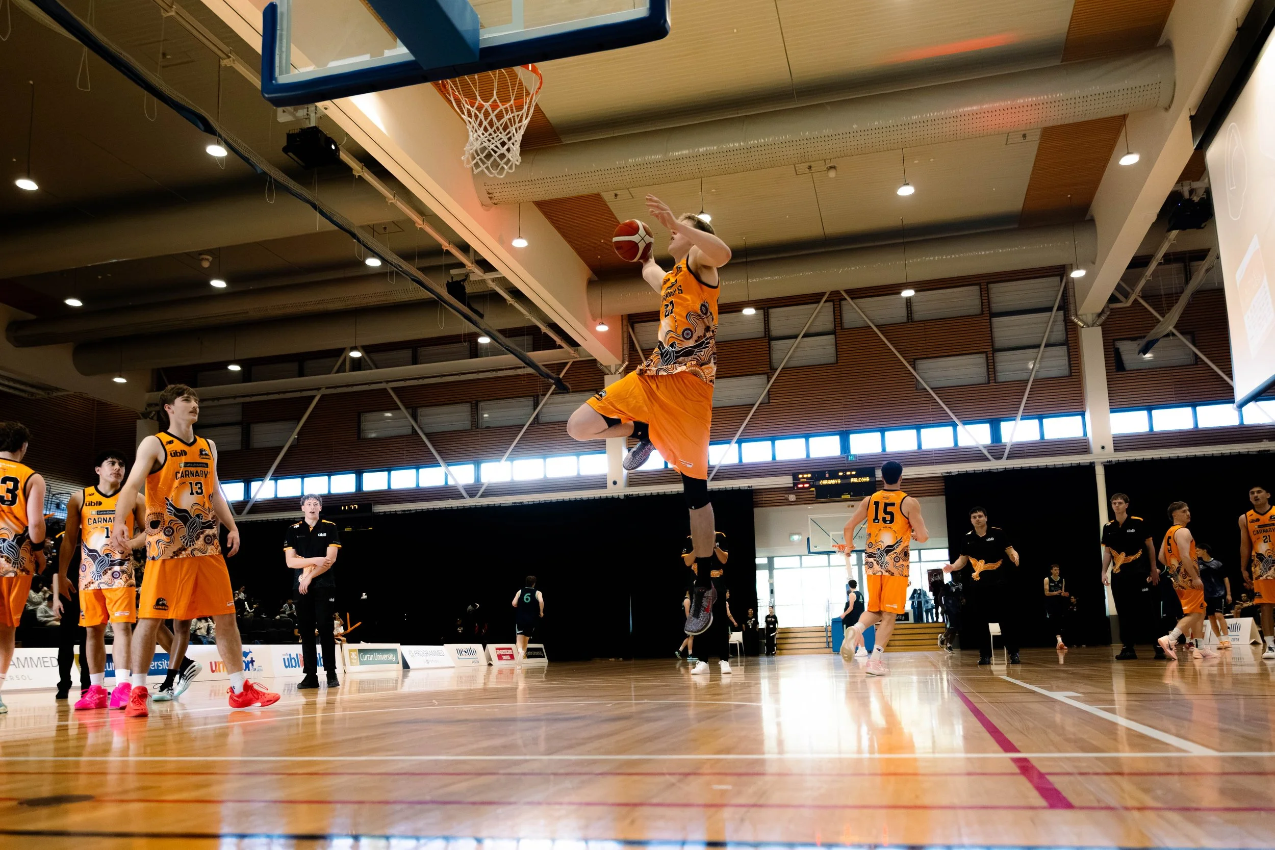 A basketball game in an indoor gym with players in orange jerseys, one jumping to catch the ball near the hoop, others watching or preparing to play.