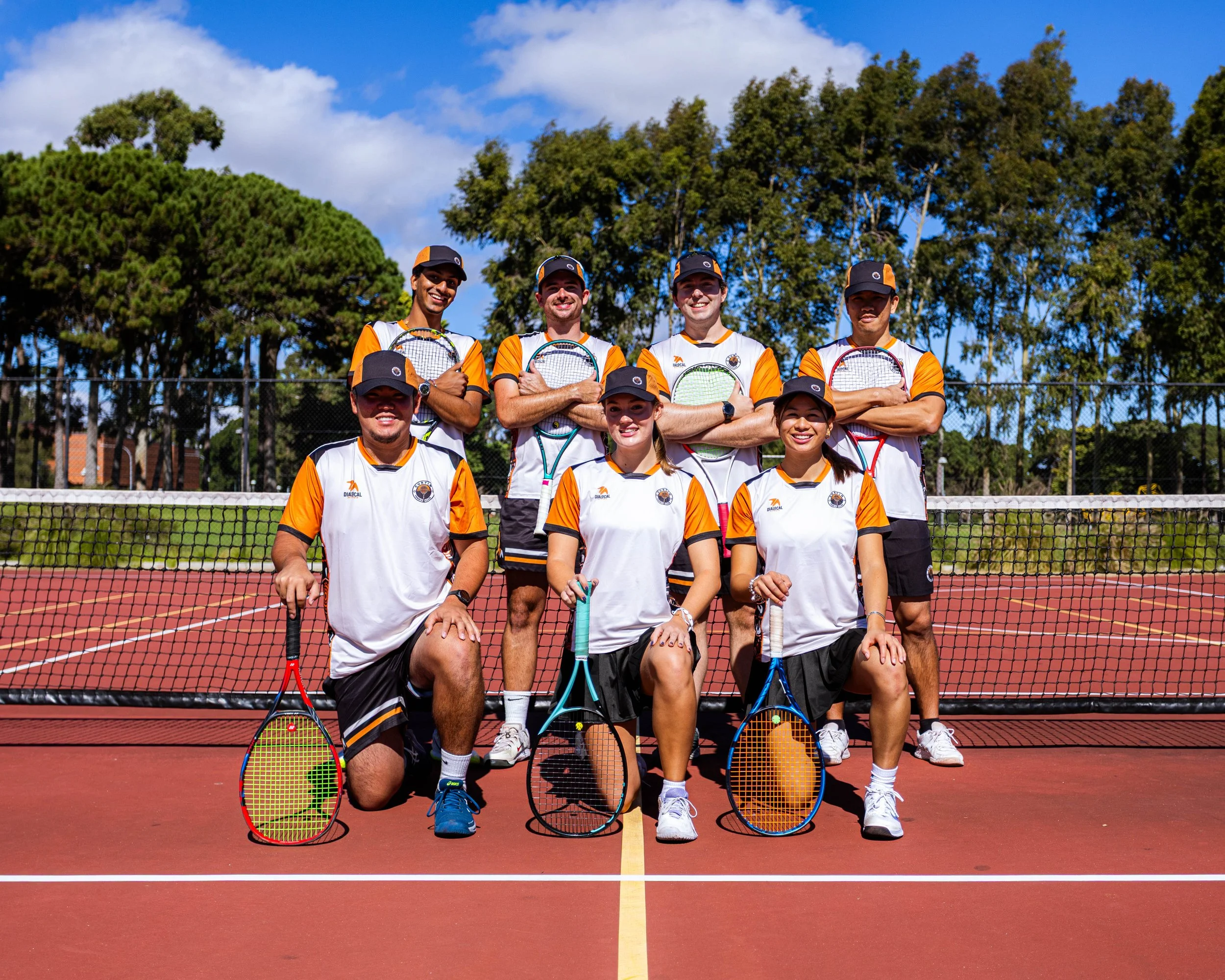 A group of eight tennis players, four men and four women, are posing on a tennis court with six tennis rackets in front of them. They are wearing matching sports uniforms in white, orange, and black, and are standing in front of a tennis net. Behind them are green trees and a partly cloudy blue sky.