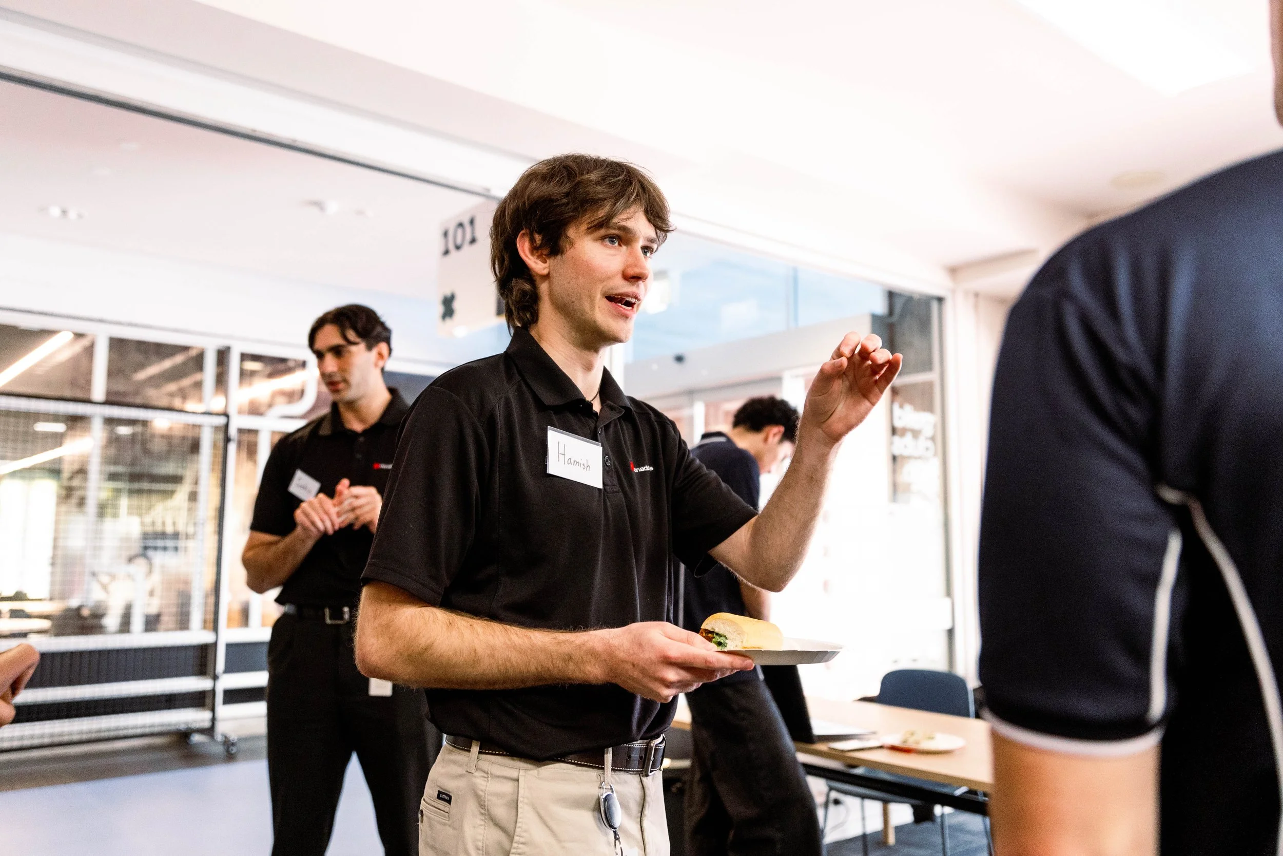 A young man with brown hair wearing a black shirt with nametag 'Hamish' is holding a plate with a sandwich, engaged in conversation at a professional or networking event.