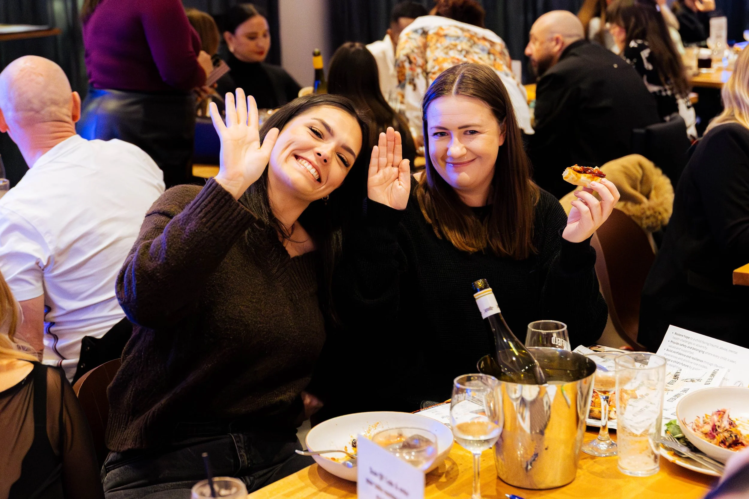Two women sitting together at a dining table smiling and waving at the camera, surrounded by plates of food and drinks in a lively restaurant or banquet setting with many people in the background.