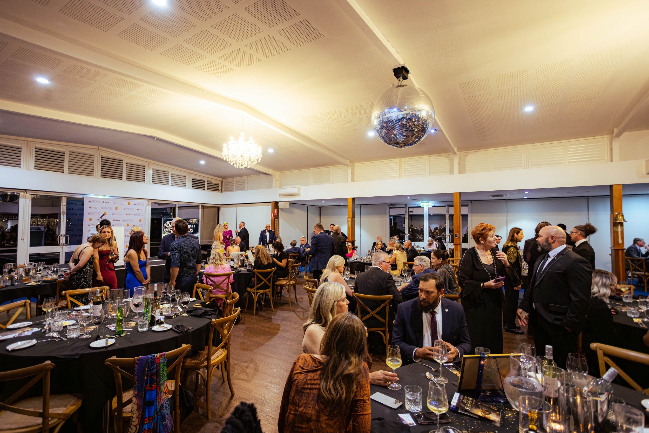 A lively banquet hall filled with people dressed in formal attire, socializing and enjoying a dinner event. The ceiling features a large disco ball and a chandelier, with tables set with glassware, plates, and silverware.
