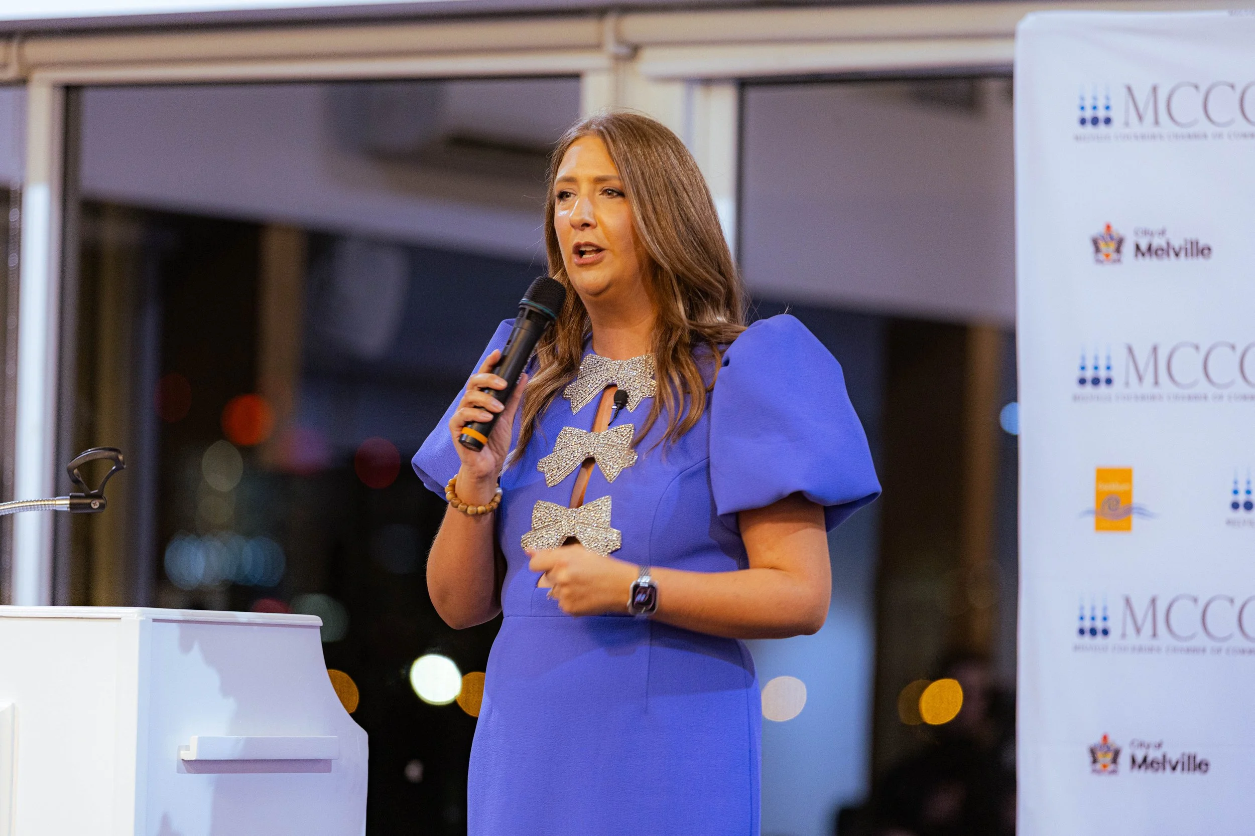 Woman with long brown hair, wearing a blue dress with large sparkly bow details, speaking into a microphone at an event with MCCG logo in the background.