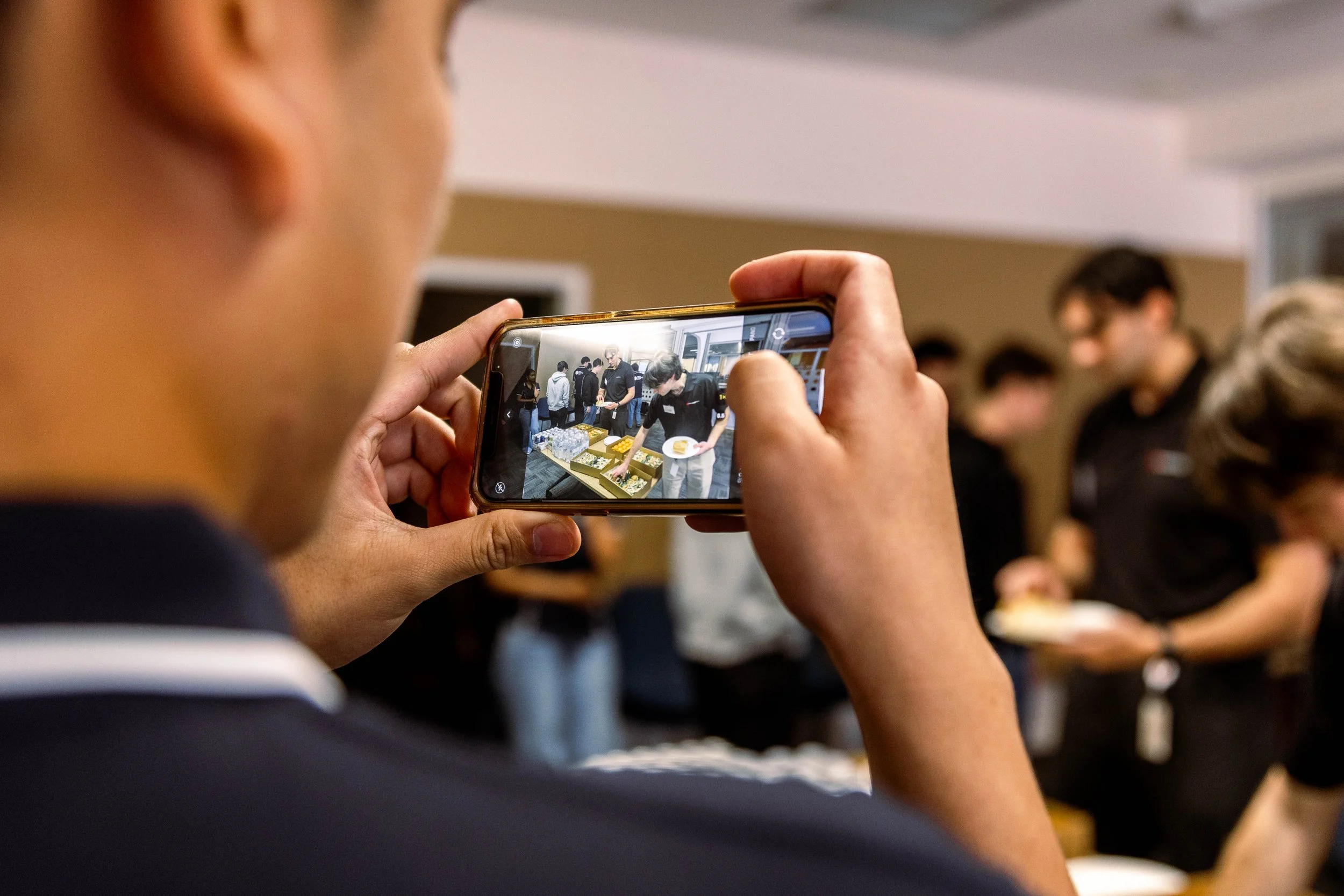 Person taking a photo of a group of people waiting in line, possibly at a food service or event.