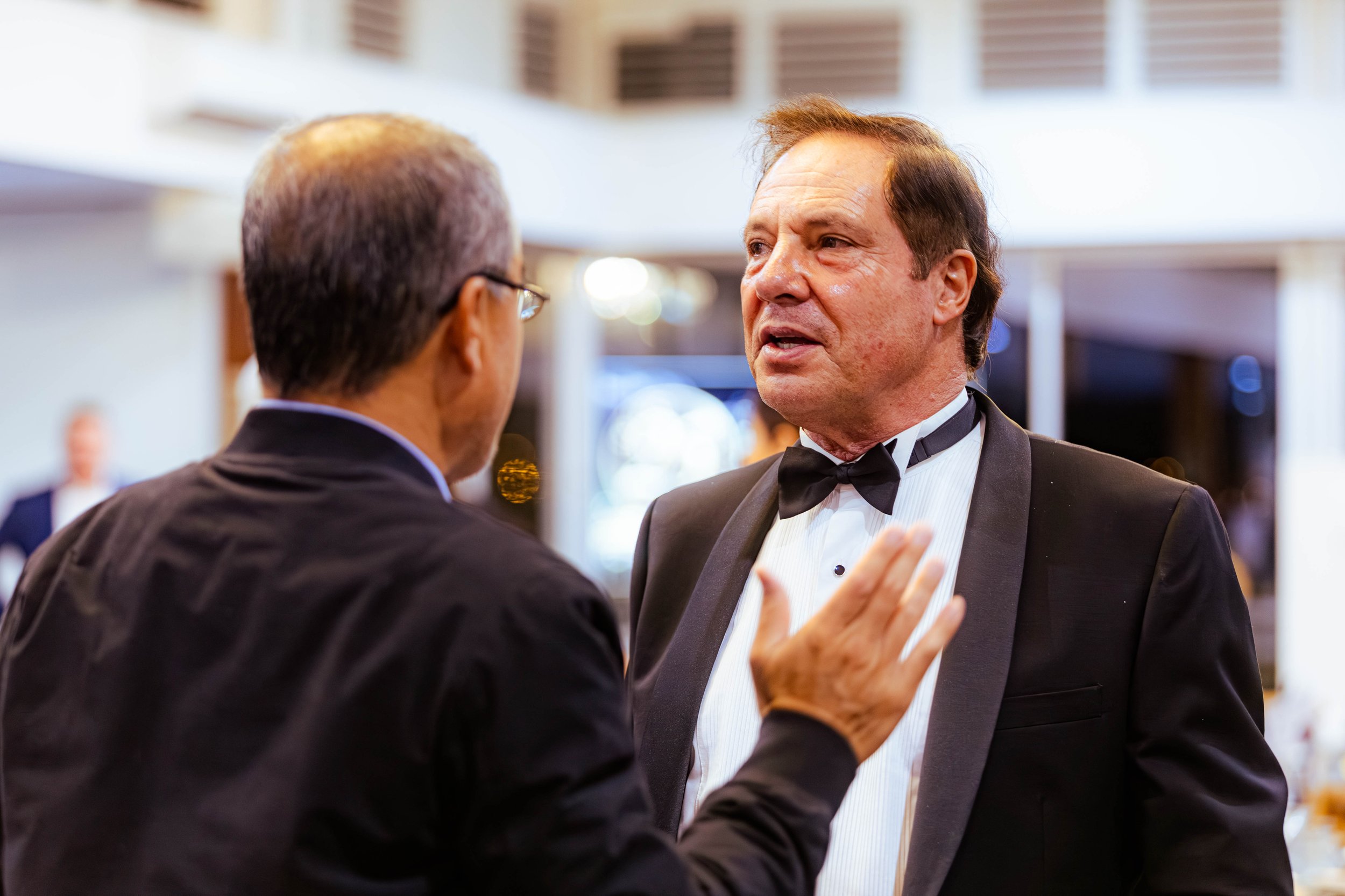 Two men in tuxedos engaged in a conversation at an indoor event.