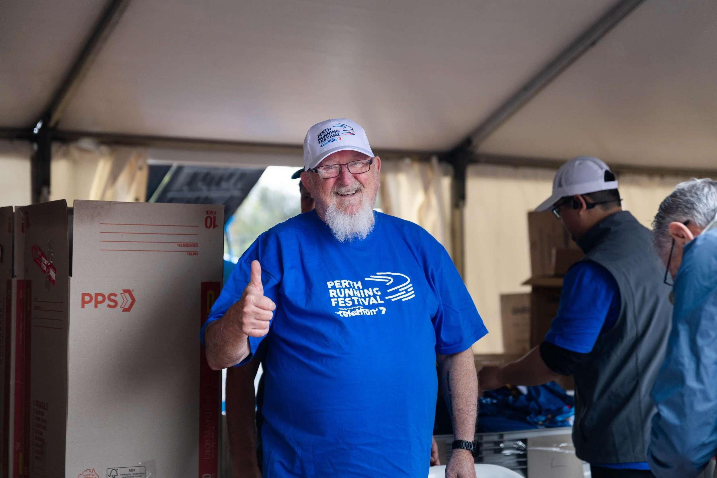 A man smiling and giving a thumbs-up, wearing a blue T-shirt and a white cap with "Perth Running Festival" logo, standing inside a tent with other people.