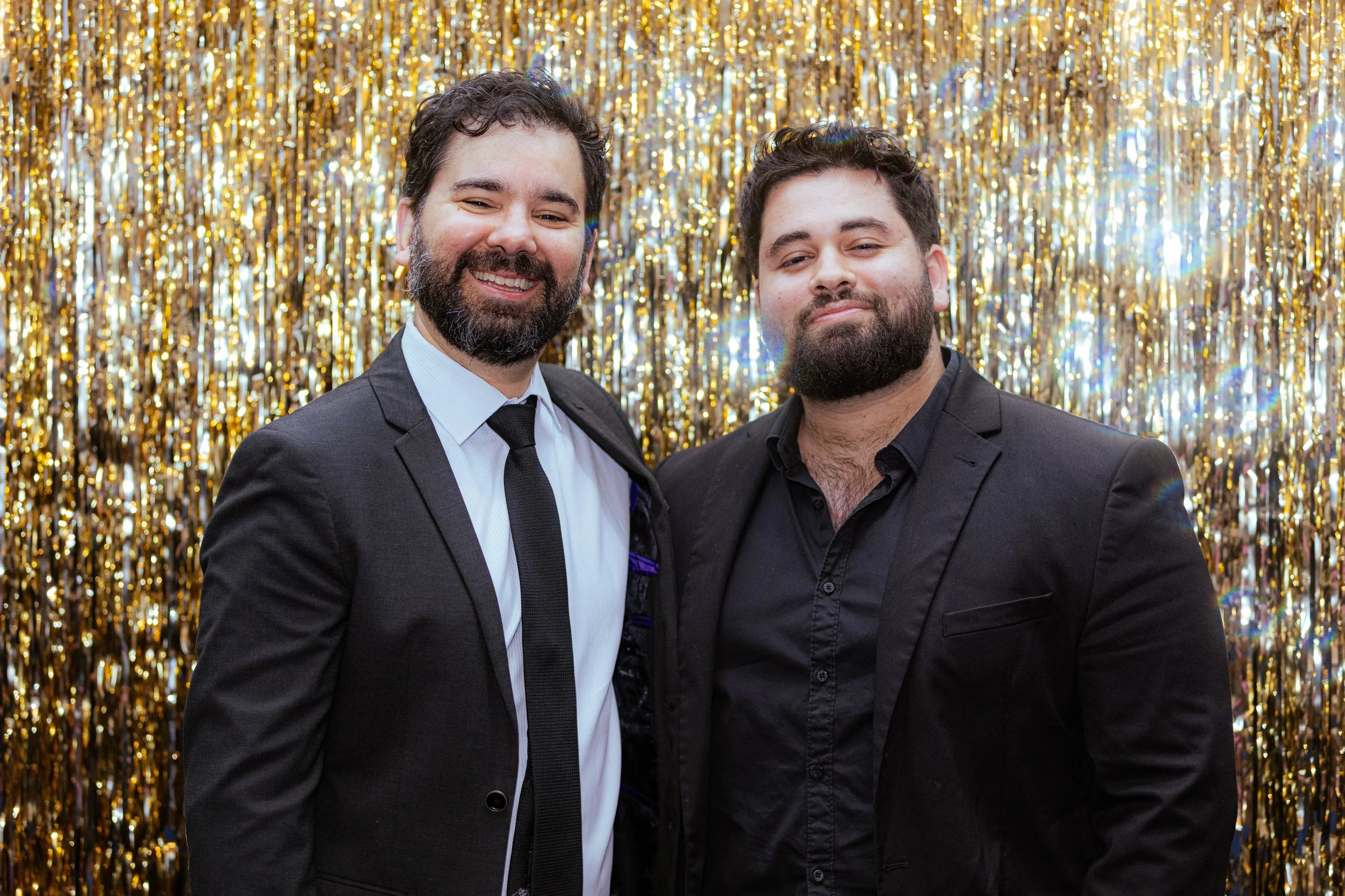 Two men in suits smiling in front of a gold, shiny, curtain background.