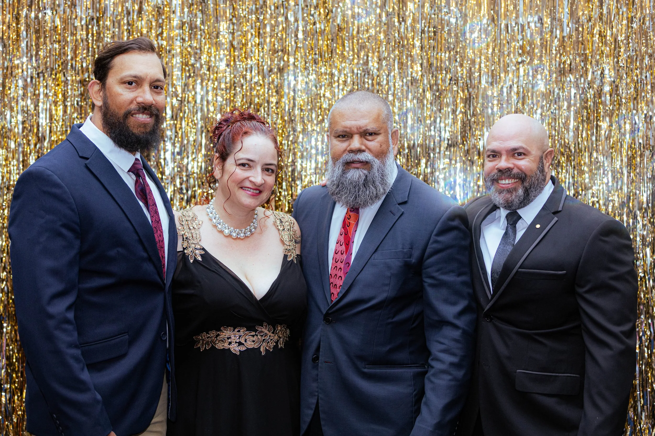 Four adults dressed in formal attire posing in front of a gold metallic fringe backdrop at a celebration or event.