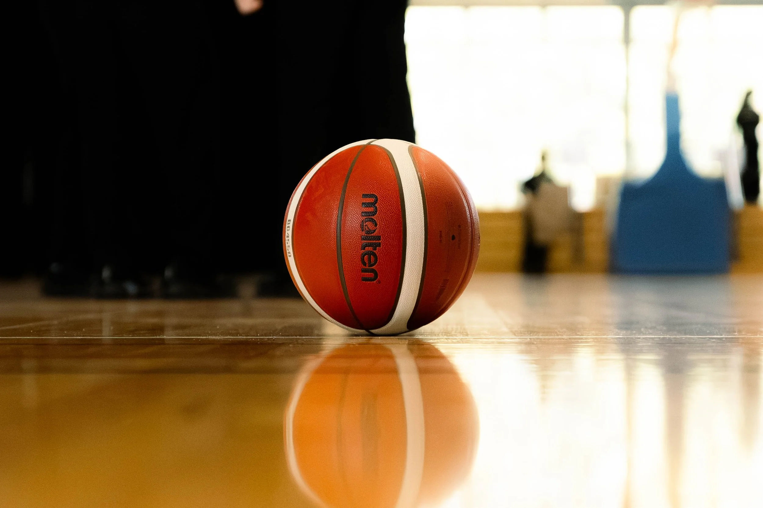Close-up of an orange and white basketball on a wooden court floor with a reflection visible, background slightly blurred with gym equipment and bright windows.