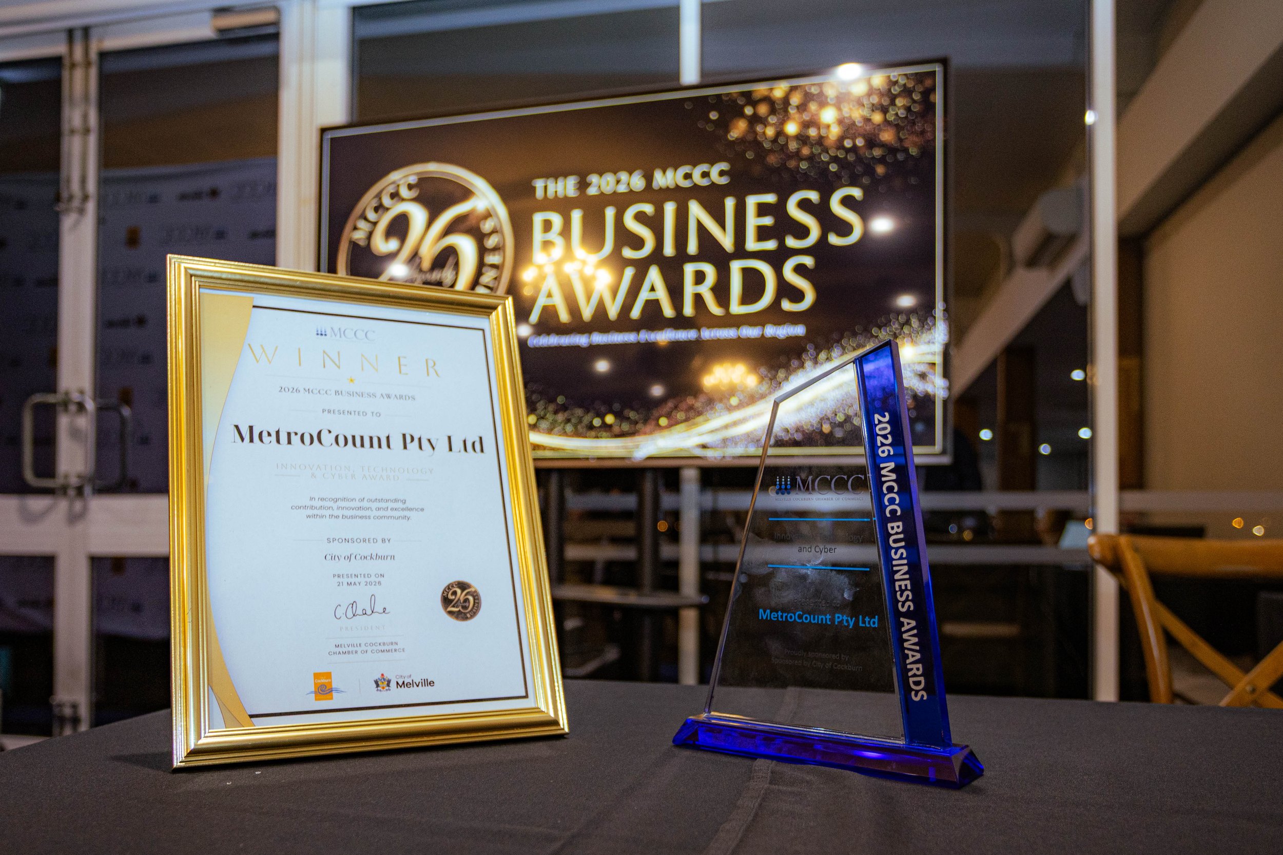 An award plaque and a glass trophy displayed on a table at the 2026 MCCC Business Awards event. The plaque is gold framed, and both items recognize achievements by MetroCount Pty Ltd. In the background, a large digital screen displays the event detai