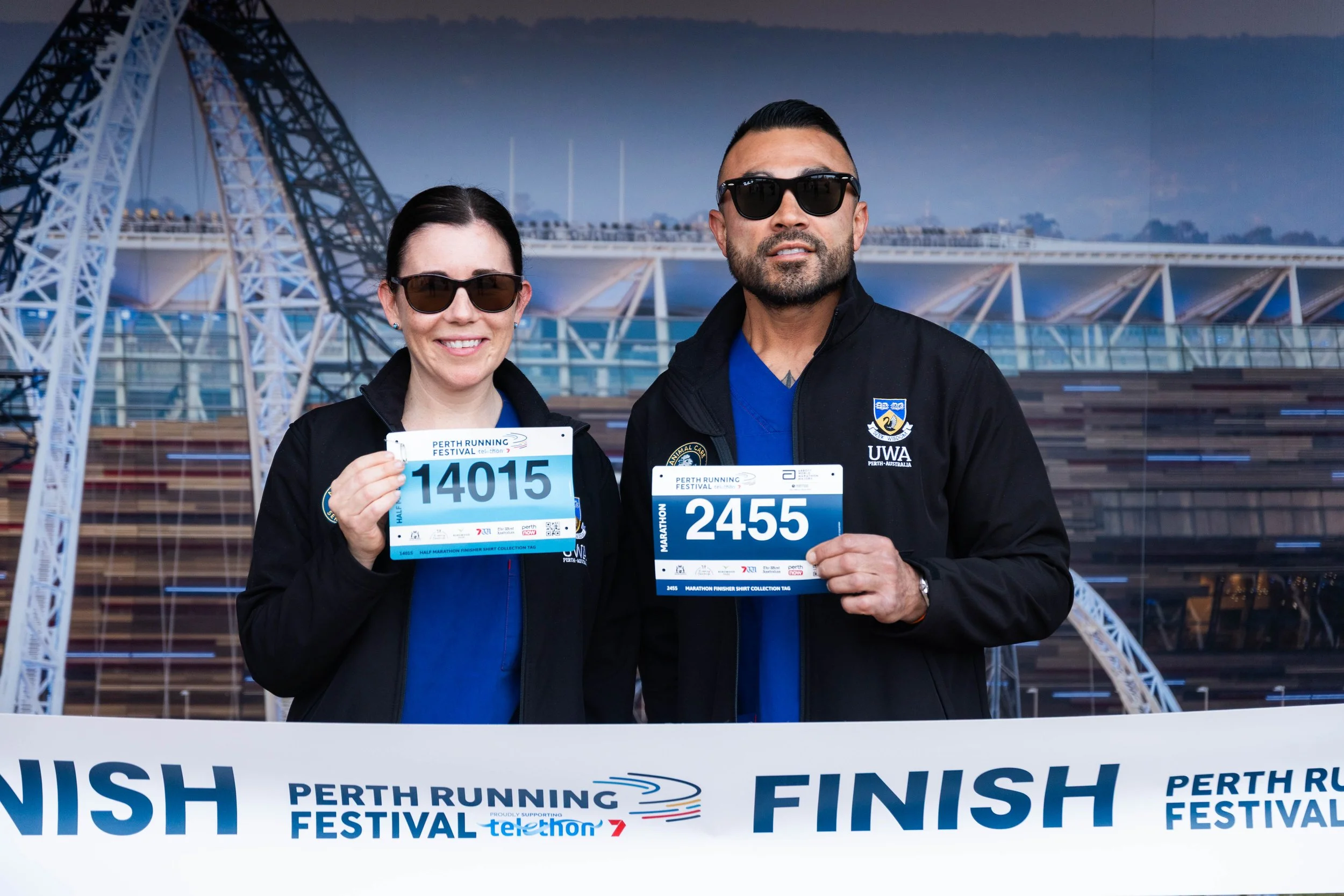 Two runners holding marathon finishing medals at the Perth Running Festival, standing in front of a backdrop showing the Perth skyline and a bridge with a large Ferris wheel.