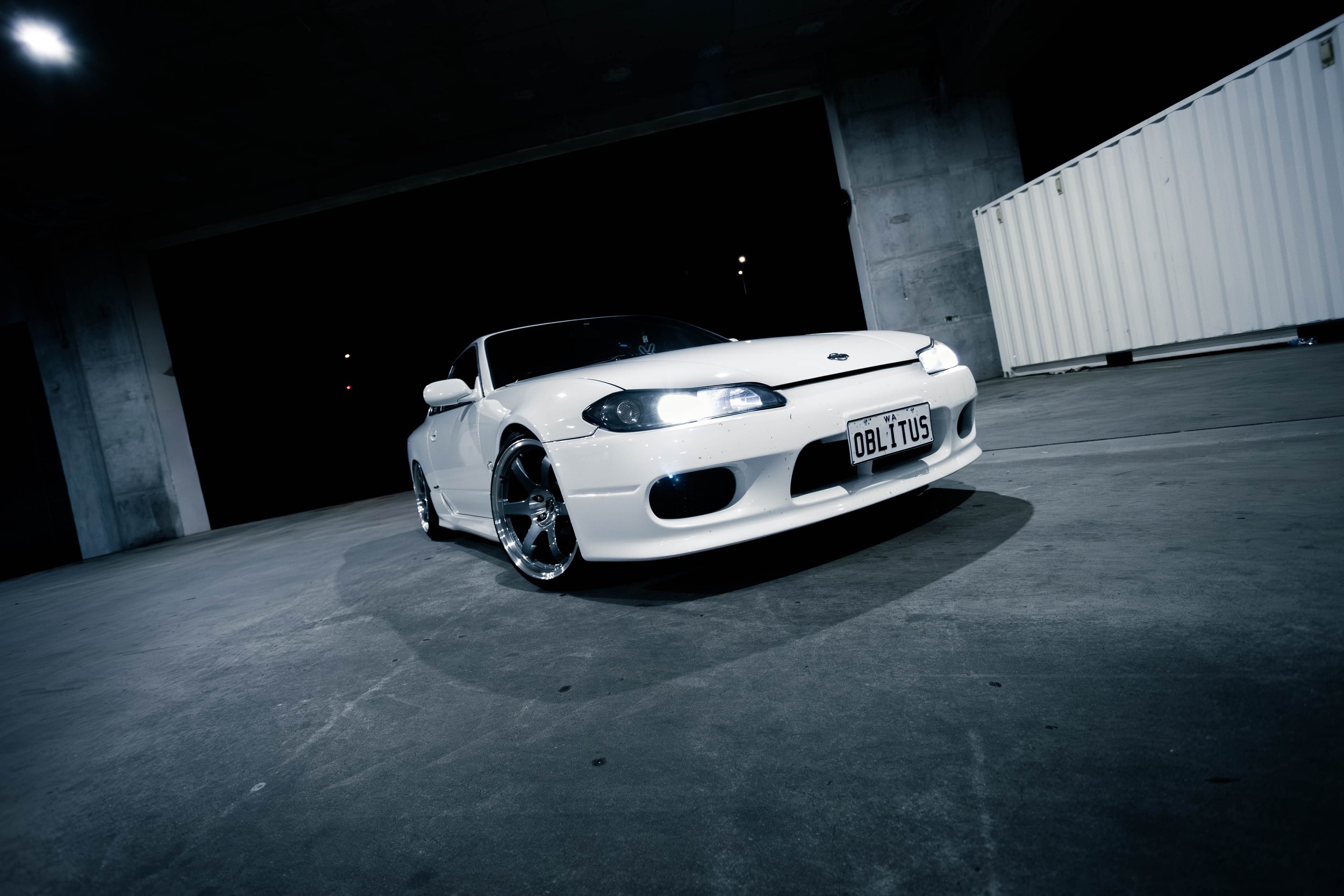 White sports car with custom license plate 'OBLITUS' parked in a dimly lit underground parking garage.