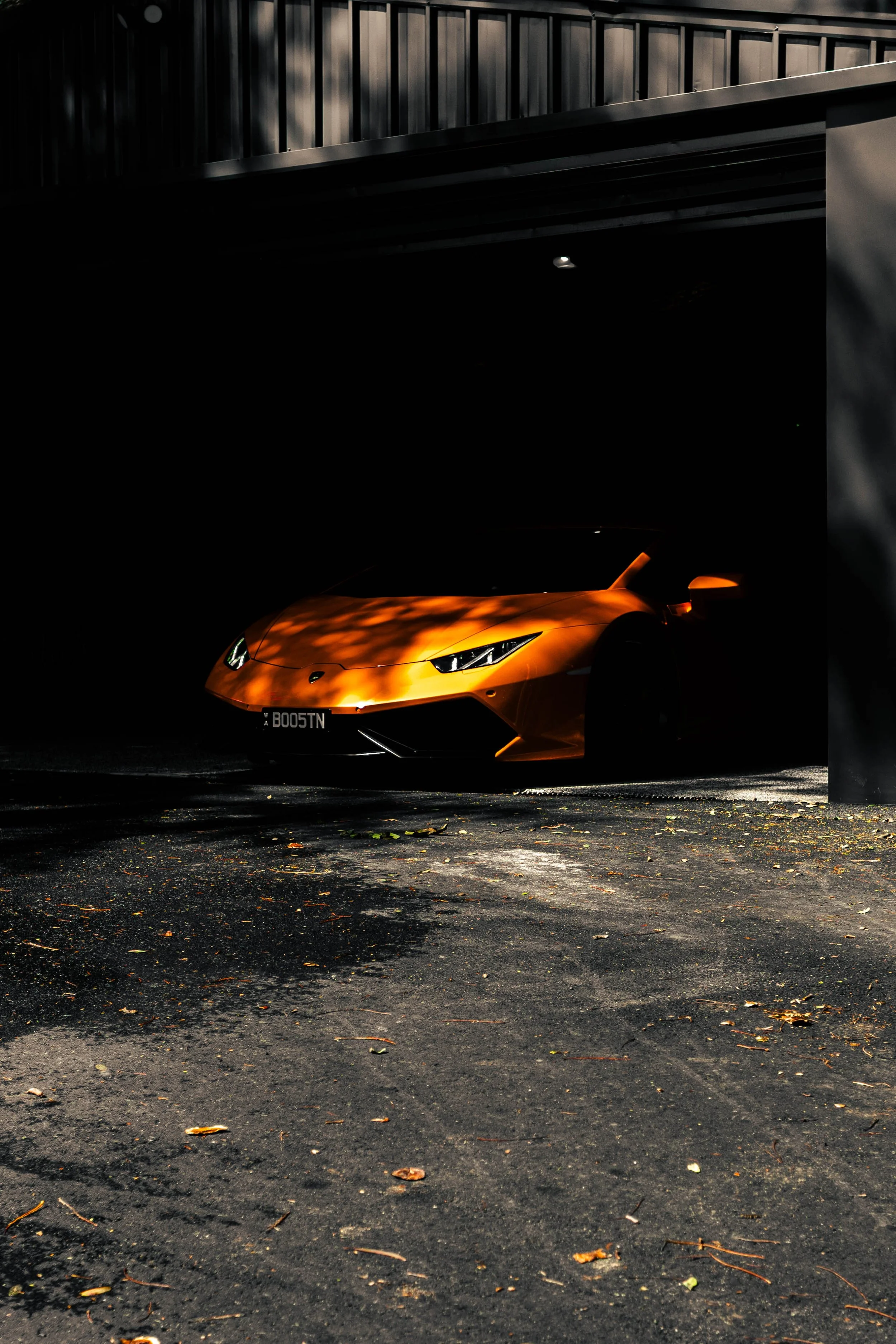 A yellow sports car, possibly a Lamborghini, parked under a structure with a dark background, partially obscured by shadows and a column in the foreground, with a license plate reading 'B00STN'.