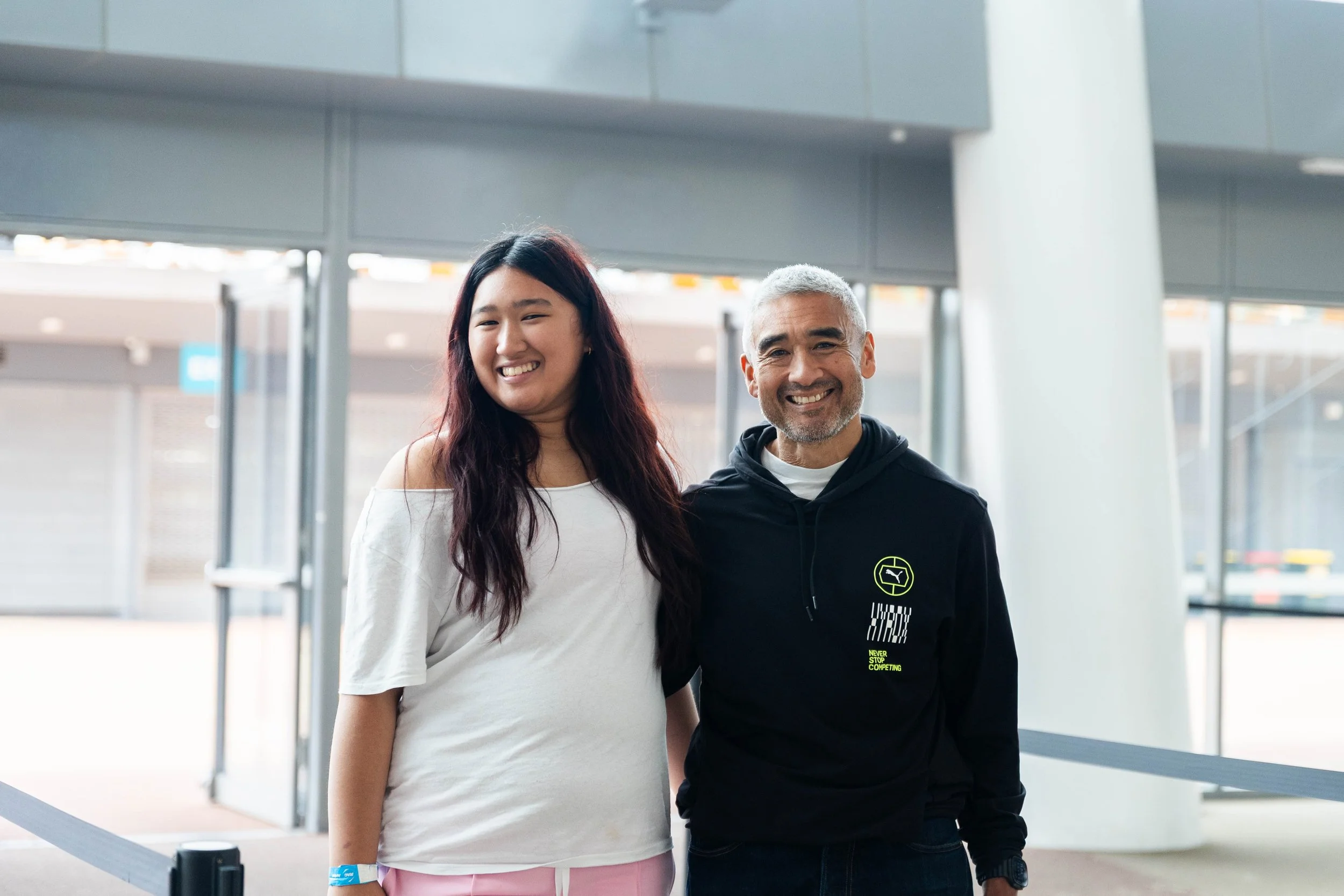 A young woman with long dark hair and an older man with short gray hair are smiling and standing close together in an indoor public space, possibly an airport or train station, with modern architecture and large windows behind them.
