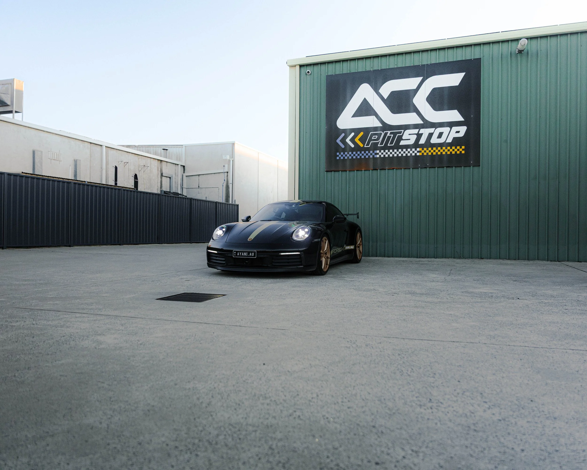 A black sports car with gold accents parked outside a building with a large ACC Pitstop sign on the green wall.